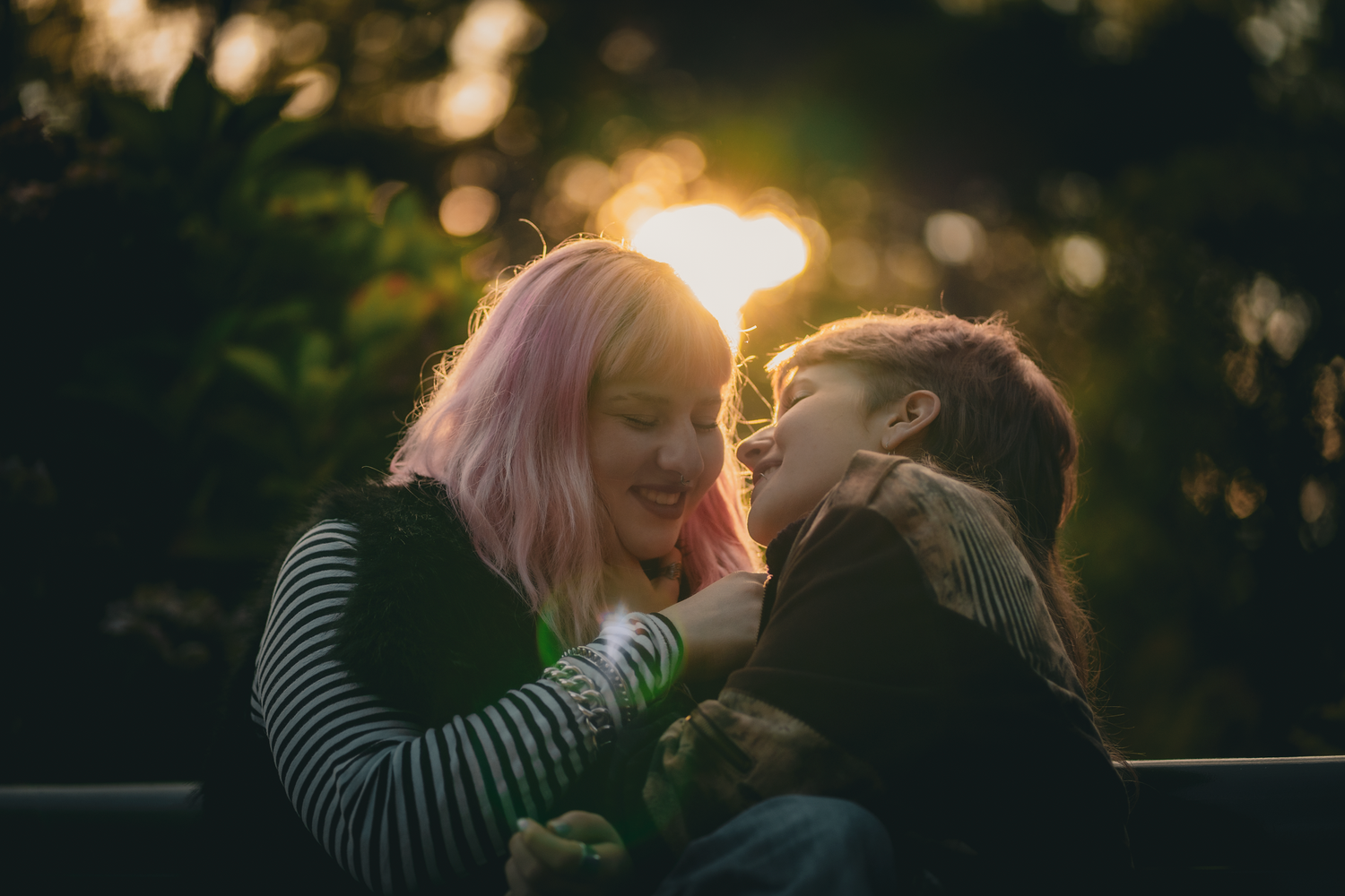 A woman with pink hair and a child share a joyful moment at sunset, laughing together outdoors.