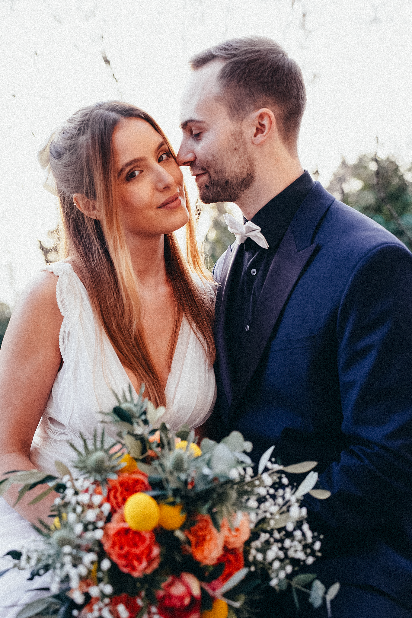 Bride and groom share an intimate moment, she holds a vibrant bouquet of orange, red, and yellow flowers.