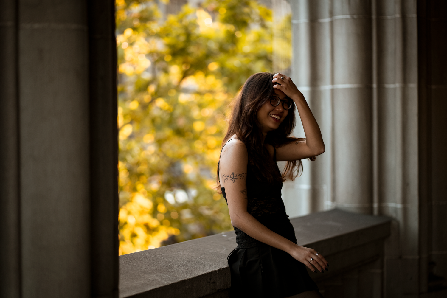 Young woman in black outfit smiling while sitting on stone ledge with autumn foliage in background.
