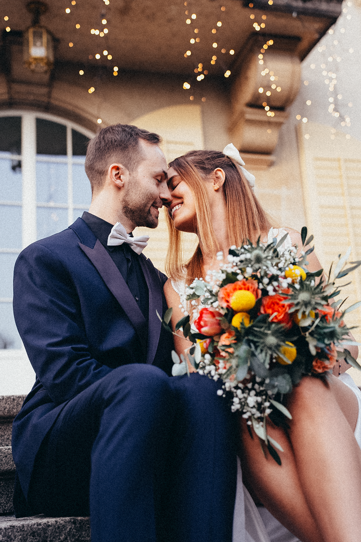 Romantic bride and groom touching foreheads on steps with bokeh fairy lights glowing in the background.