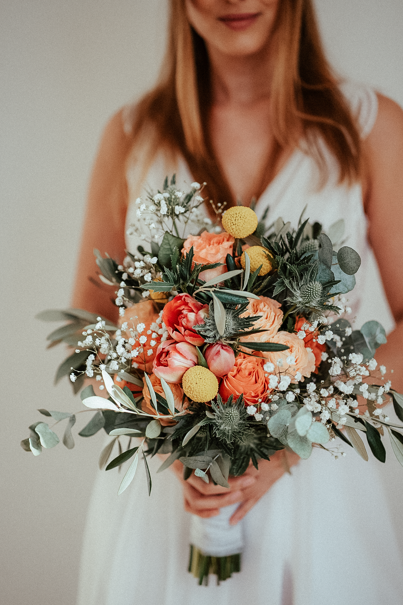 Bride in white dress holding a lush wedding bouquet with coral roses, yellow craspedia, thistles, and eucalyptus.