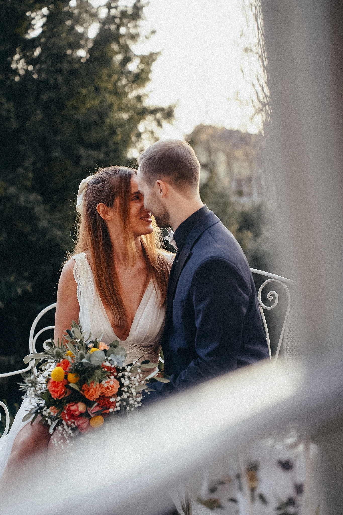 Wedding couple sitting closely on ornate white bench, sharing a romantic moment with a colorful bouquet.