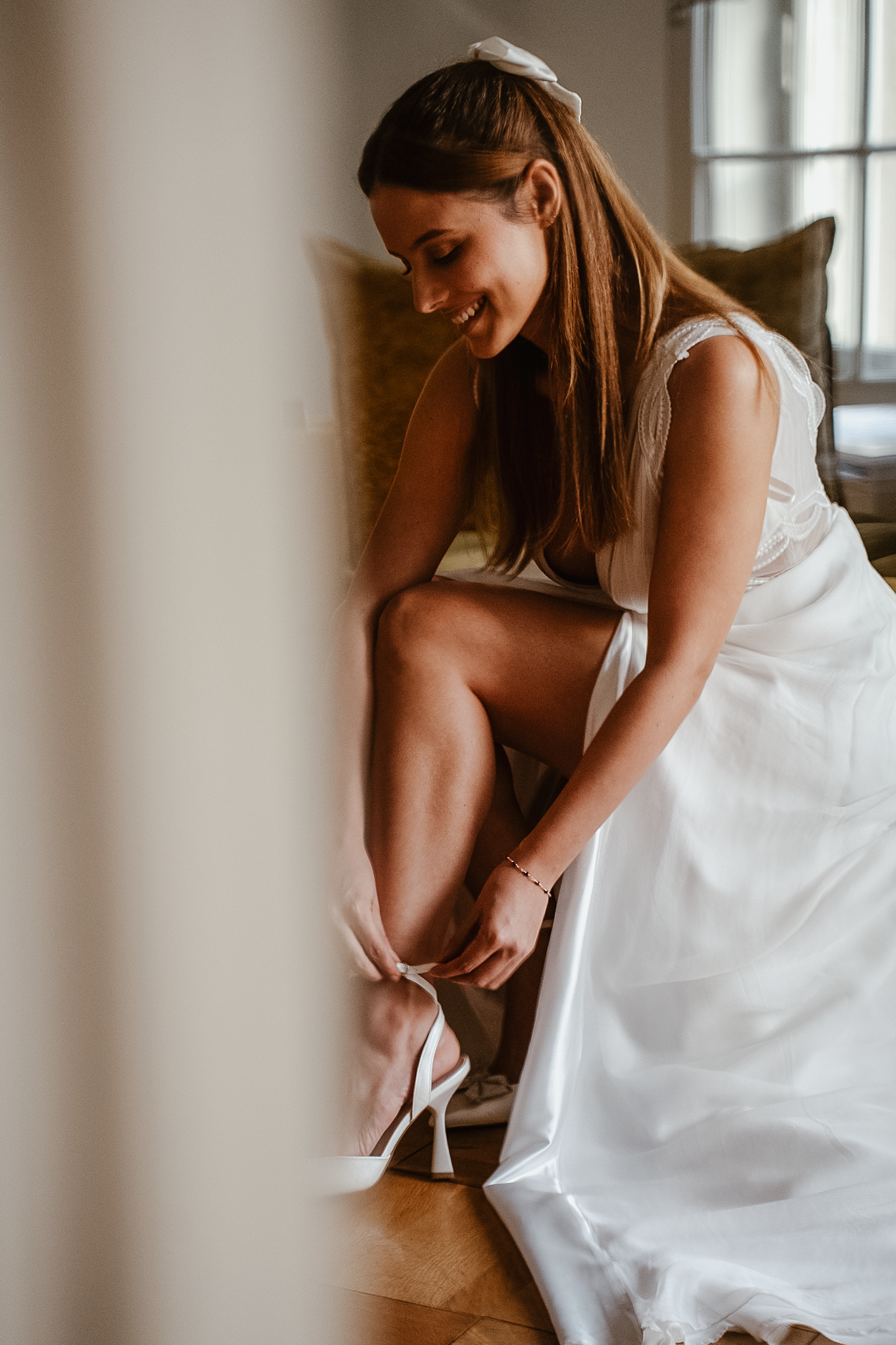 Smiling bride in white wedding dress fastening her strappy heels while seated near a bright window.