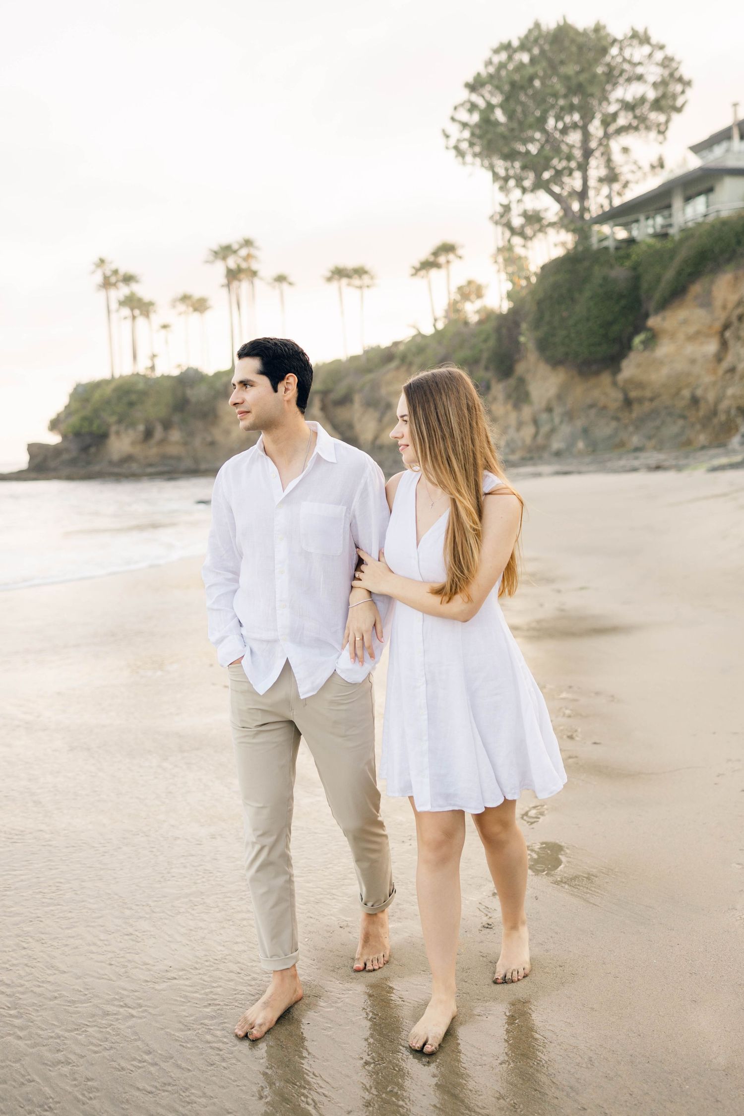 A couple in white outfits walks together on a sandy beach at sunset.