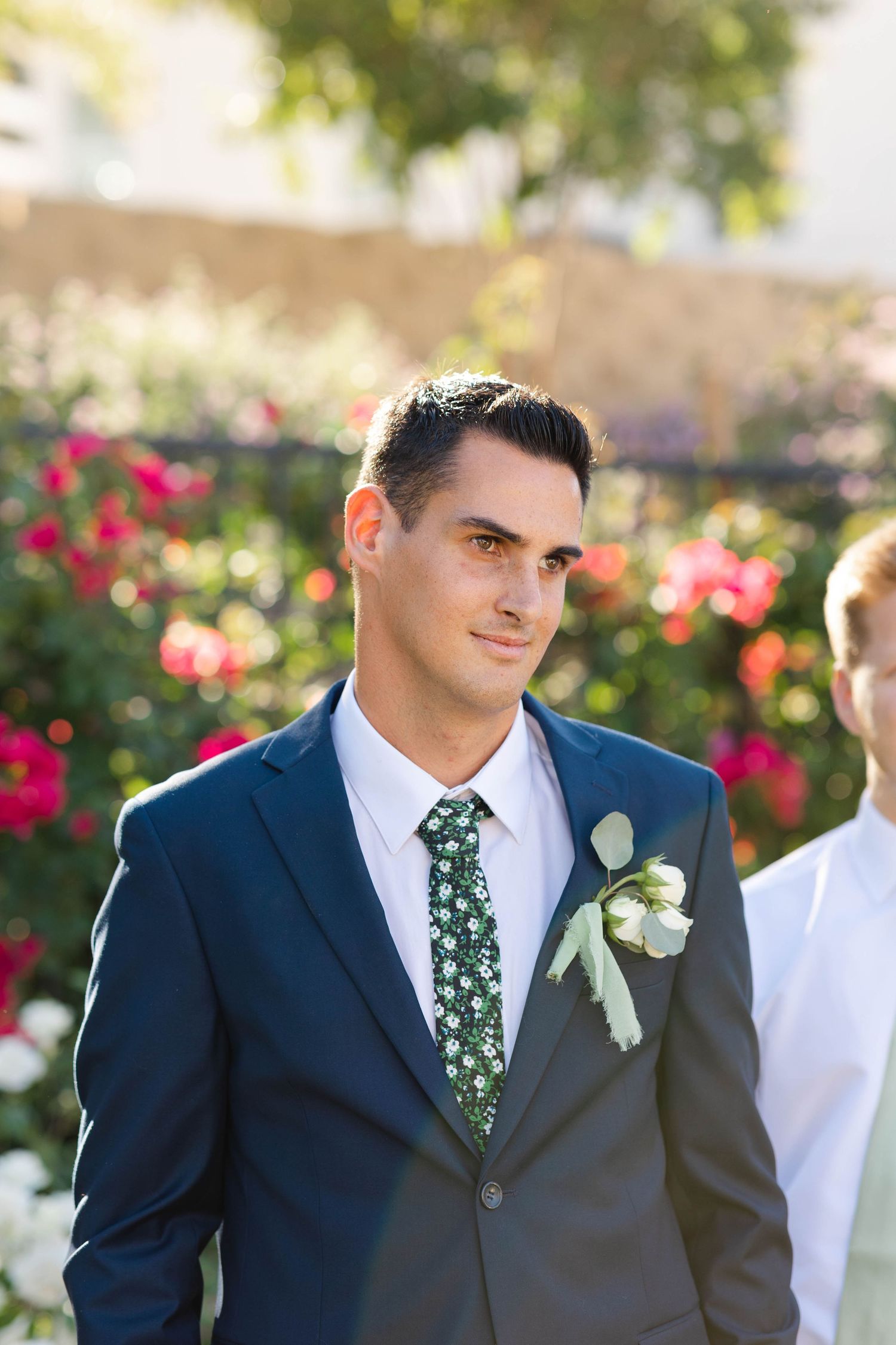 Groom in navy suit with floral tie poses during outdoor wedding ceremony.