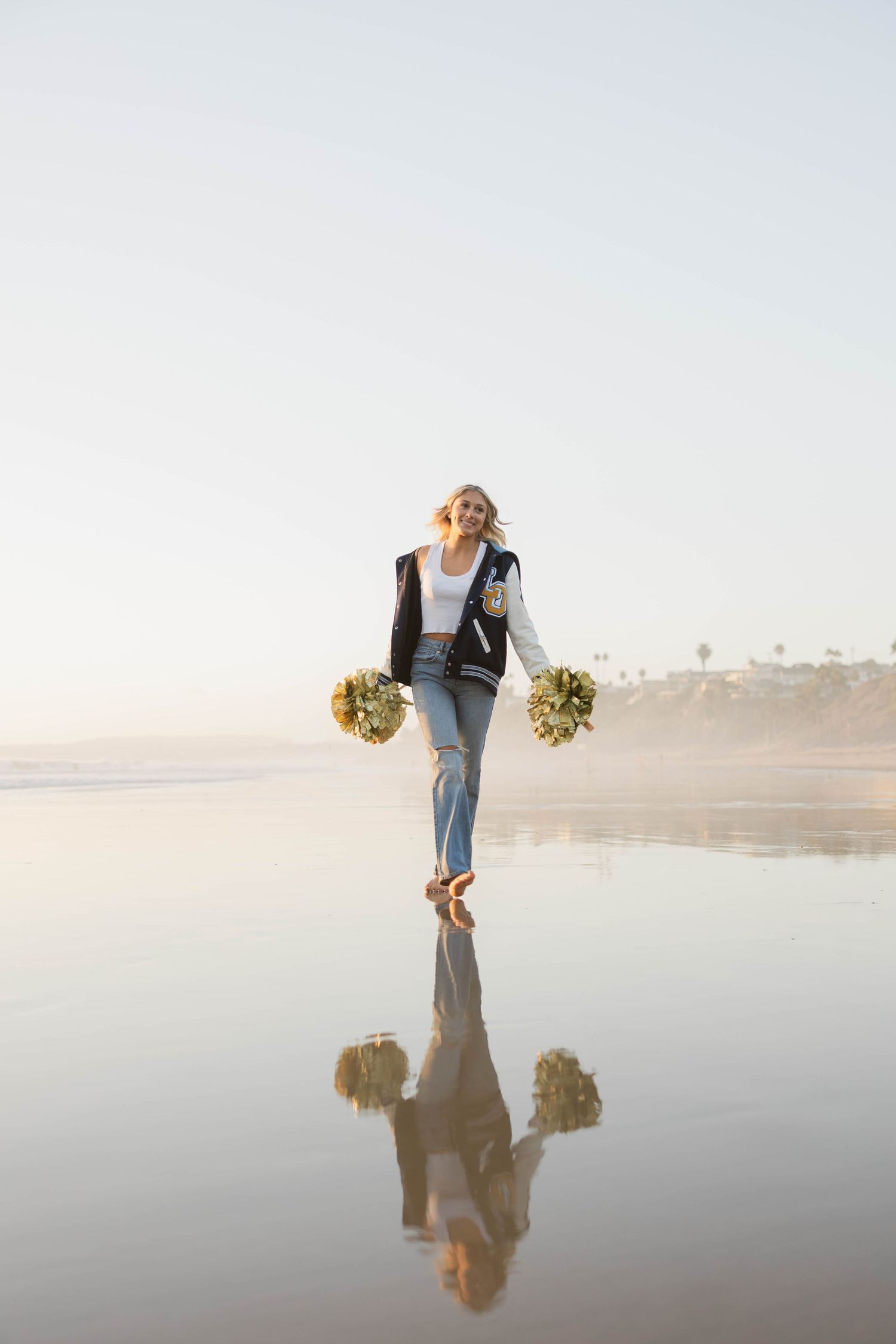 Graduate with pom poms creates reflection photos on wet beach sand.
