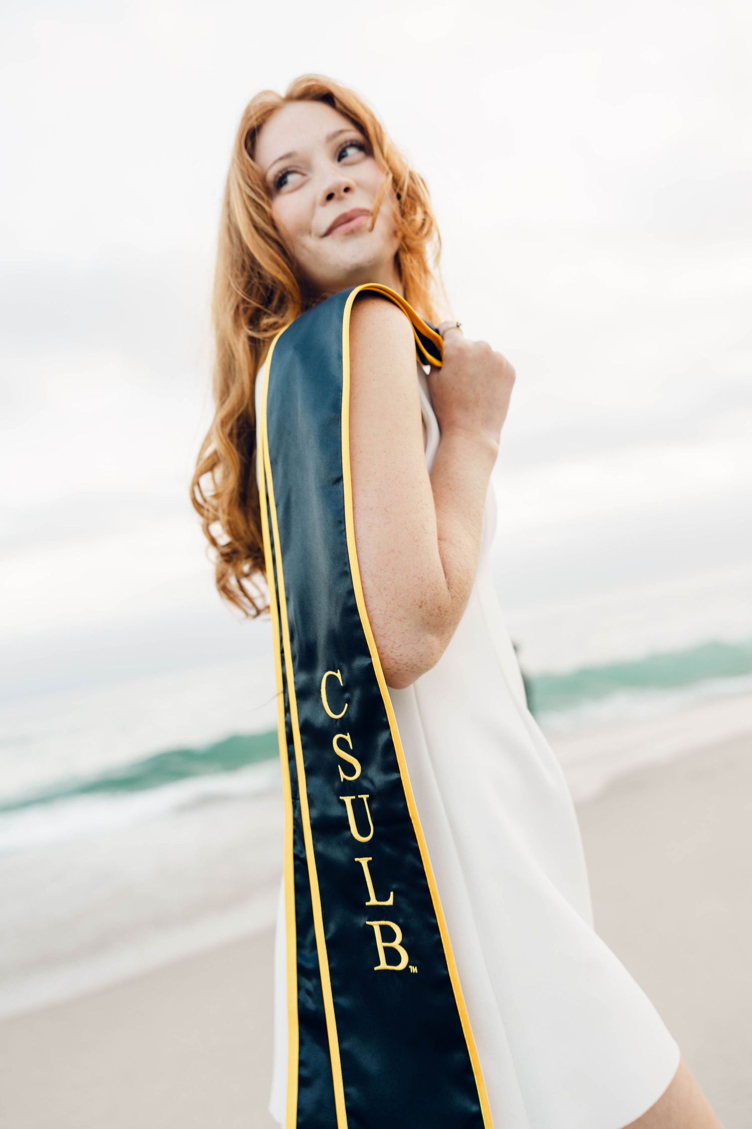 A graduate displays their graduation stole against an ocean backdrop.