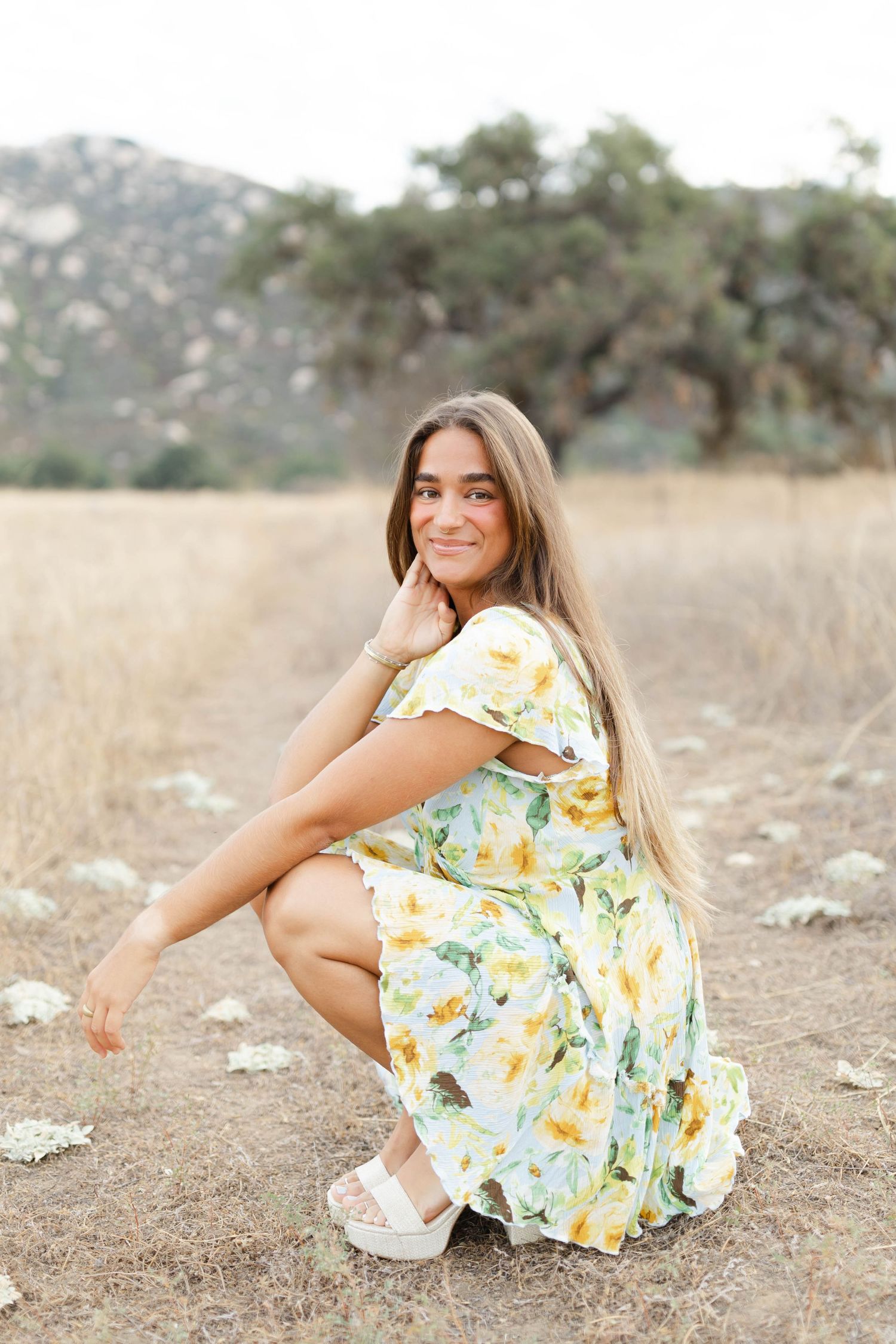 Person in floral yellow dress lying on dry grass field during golden hour photography session.