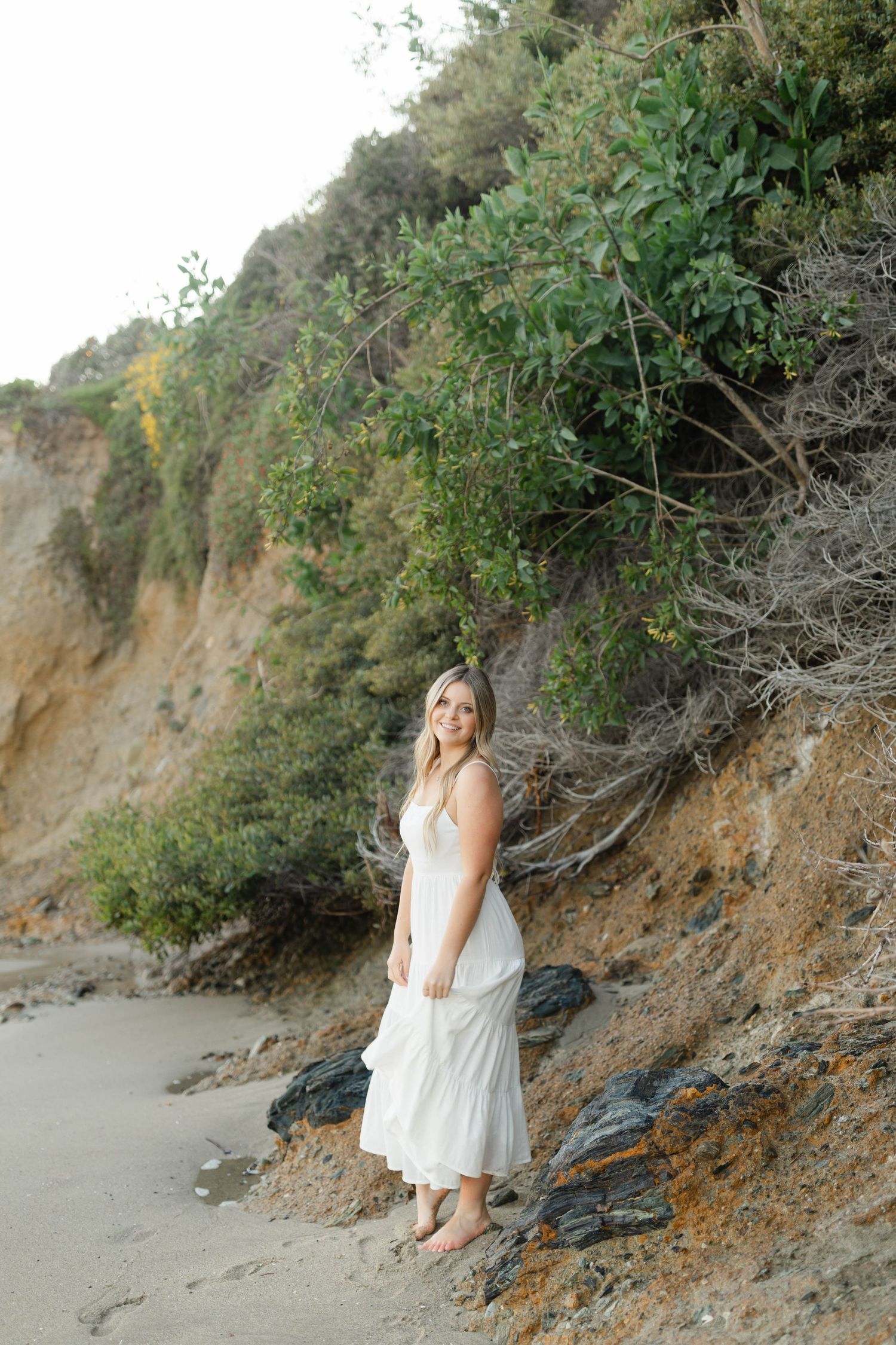 A woman in white dress stands against a coastal cliff with greenery.