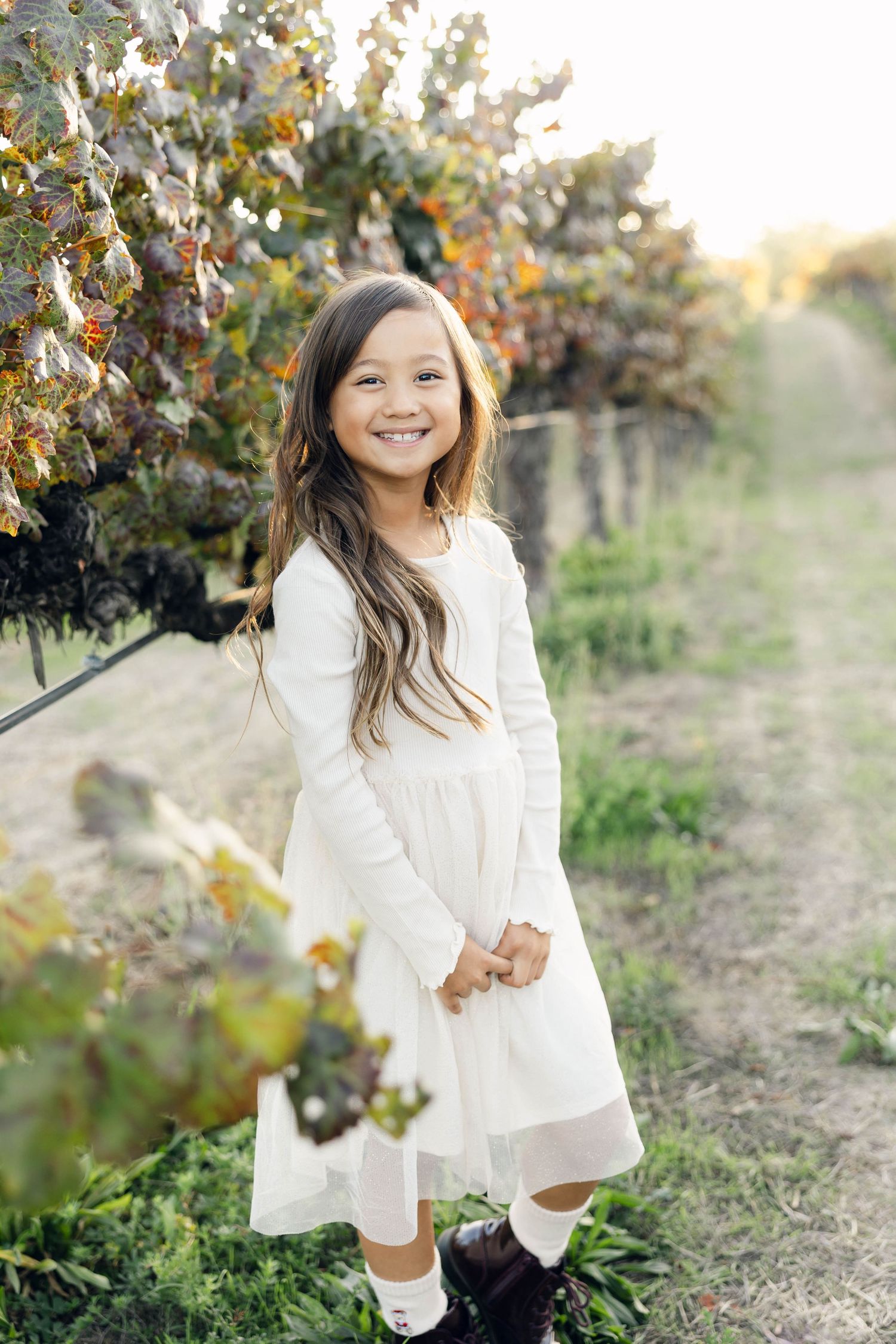 Girl in white dress playing and smiling in vineyard during golden hour.