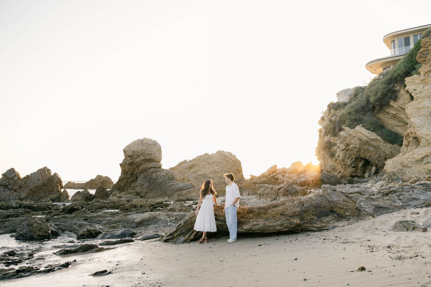 Two people in white outfits standing on rocky coastal beach during golden hour.