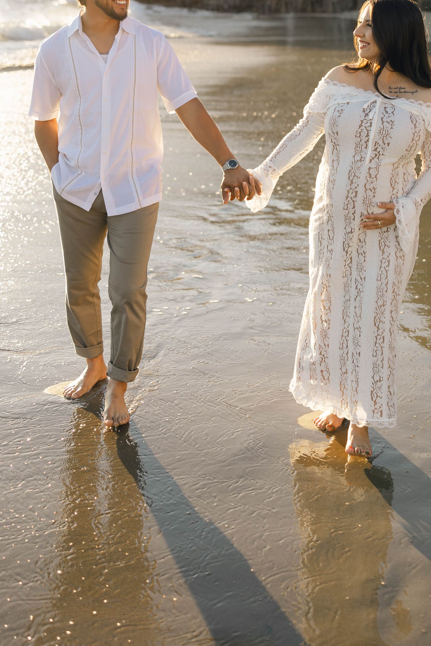 Couple holding hands while walking barefoot on wet beach sand at sunset.