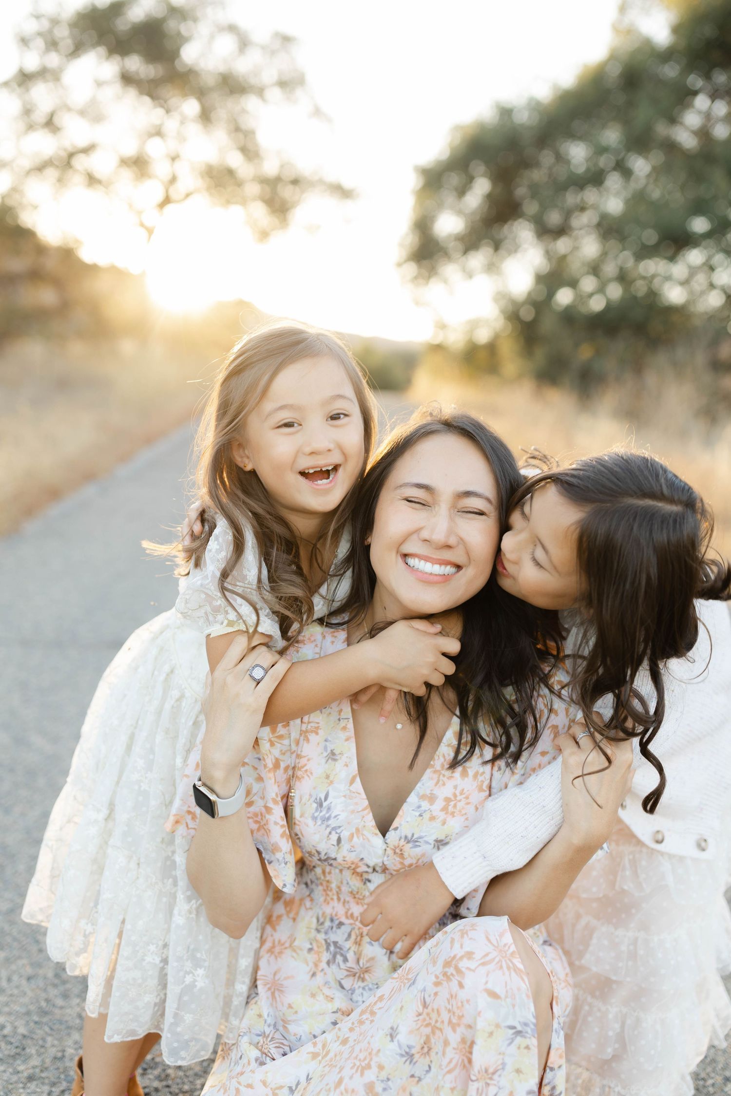 Three people in white and floral dresses sharing joyful moments during sunset outdoors.