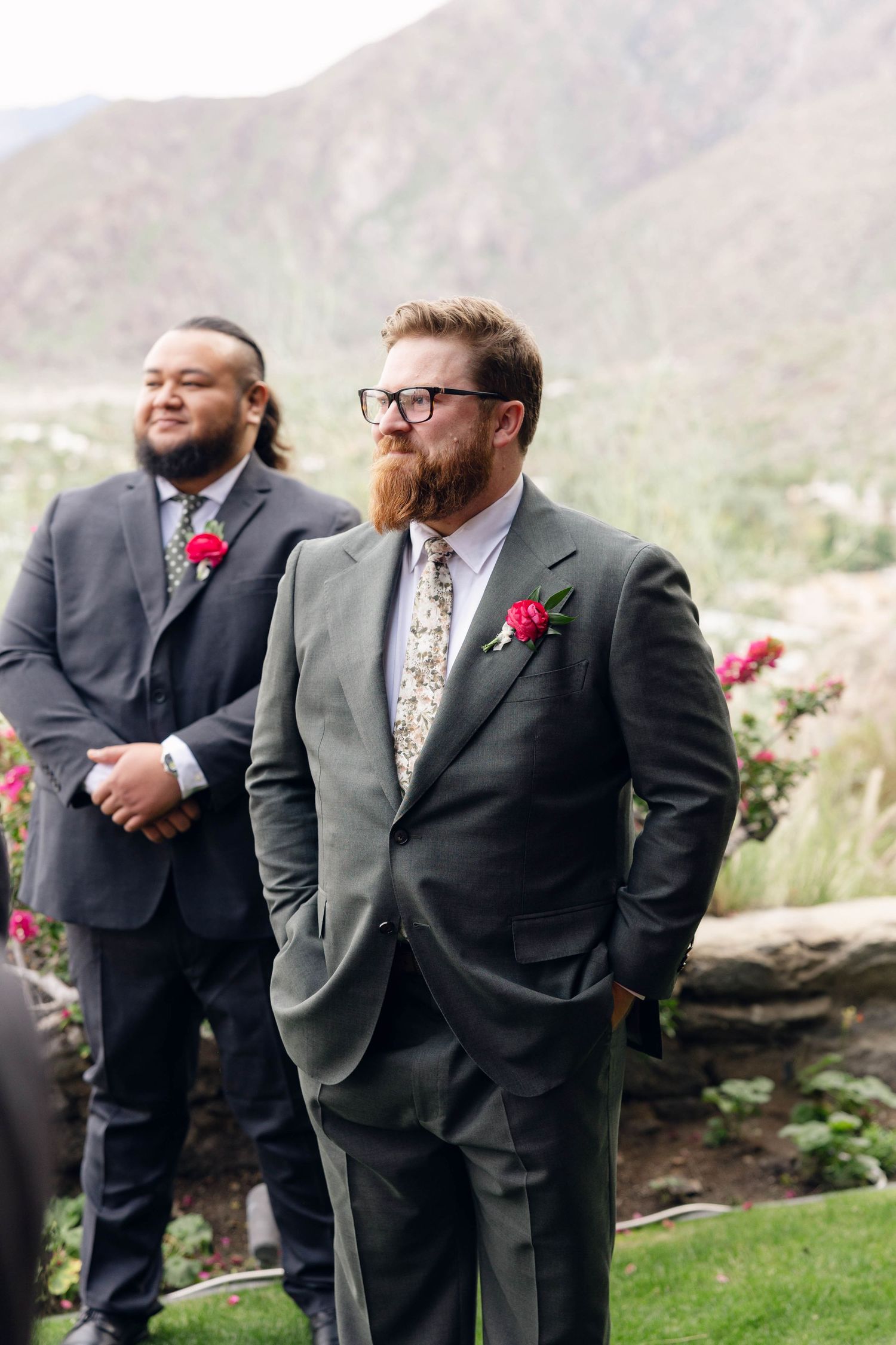 Two groomsmen in gray suits with pink boutonnieres stand together at an outdoor wedding venue with mountain backdrop.