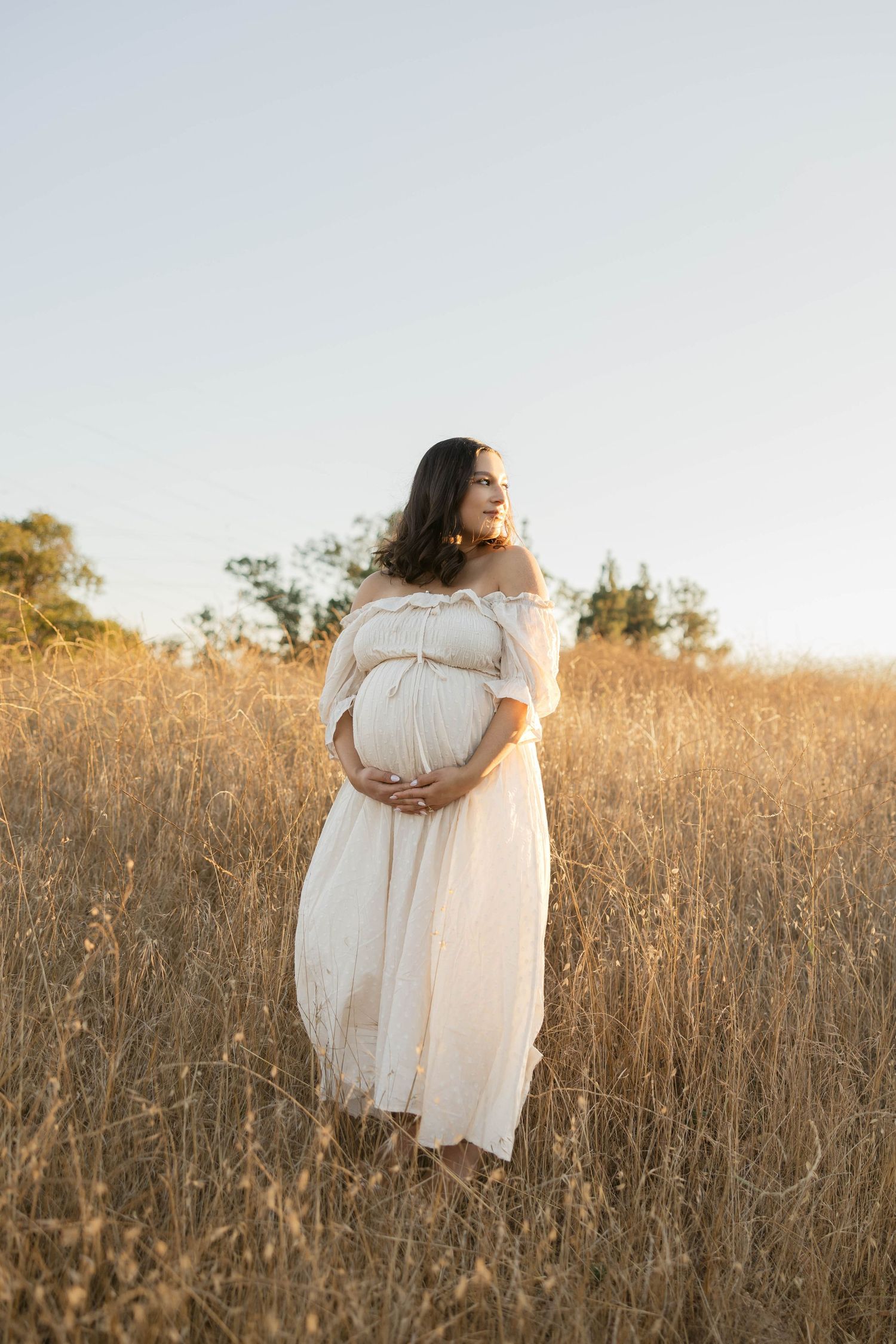 A flowing white dress contrasts against tall golden grass in a sunlit field during golden hour.