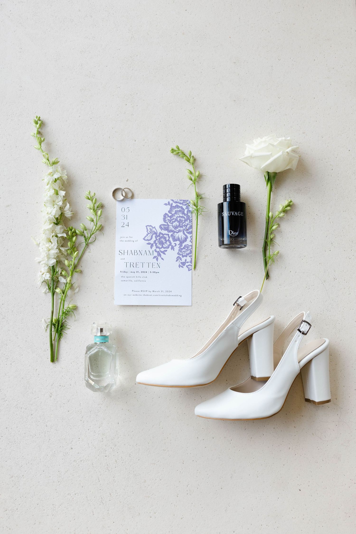 Wedding detail flatlay with white heels, flowers, and invitation on light background.