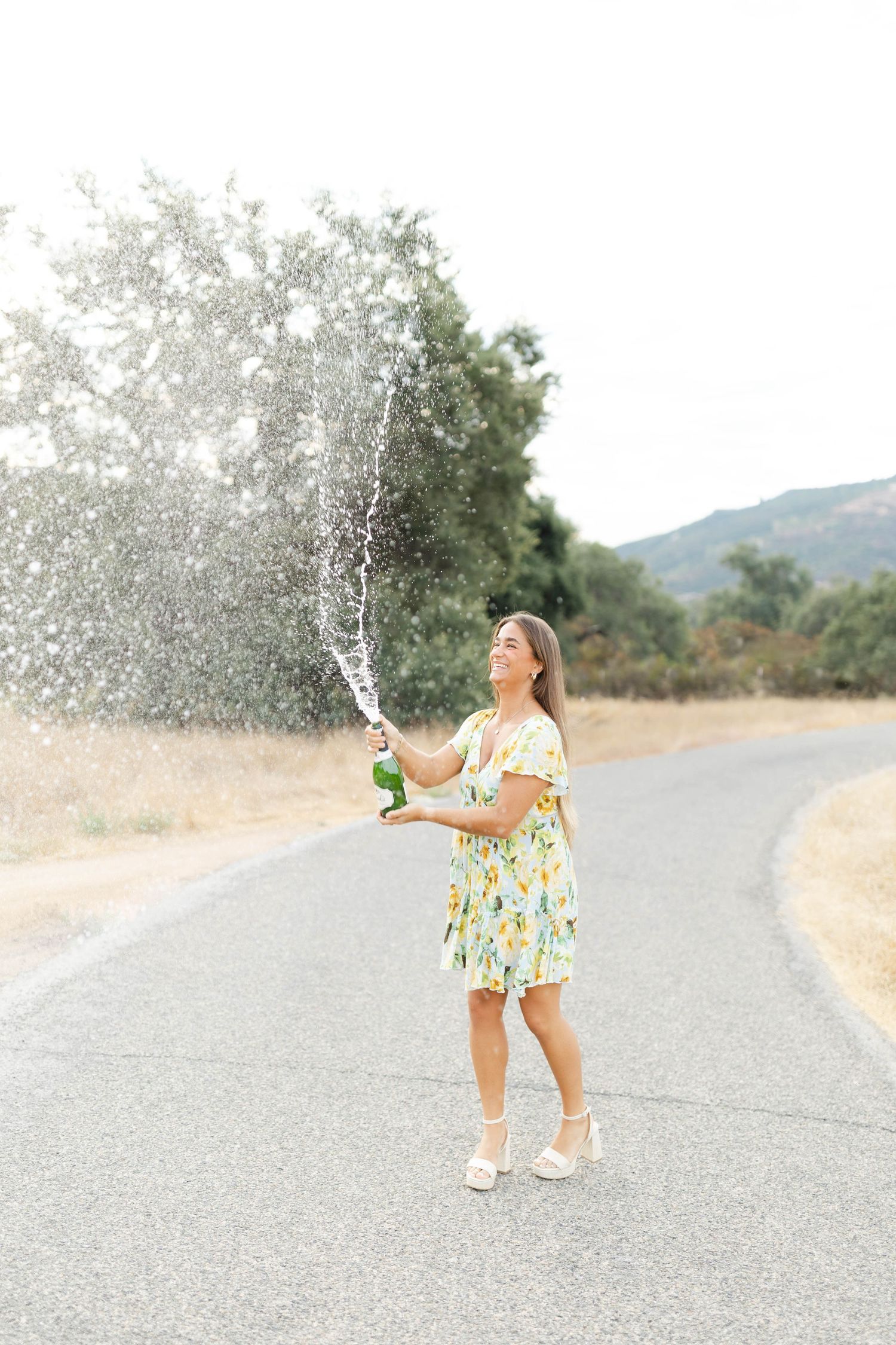 Person in floral dress spraying water from bottle on rural road with mountains in background.