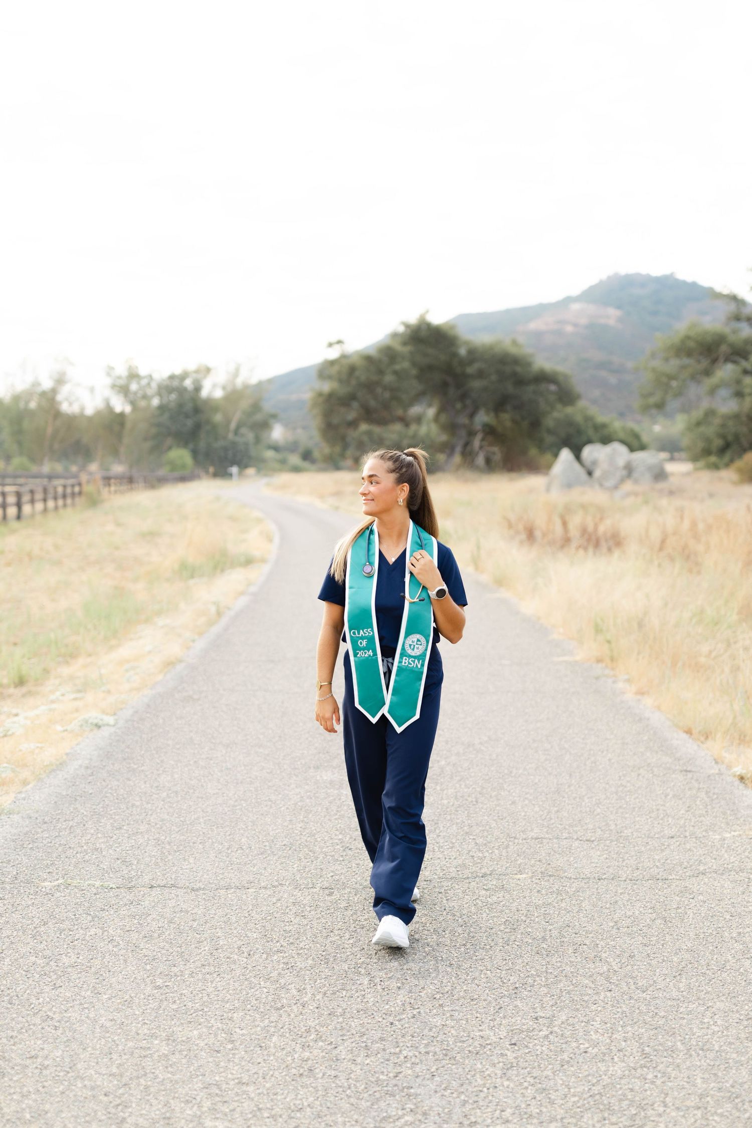 Graduate walking on rural path wearing teal graduation stole and casual attire.