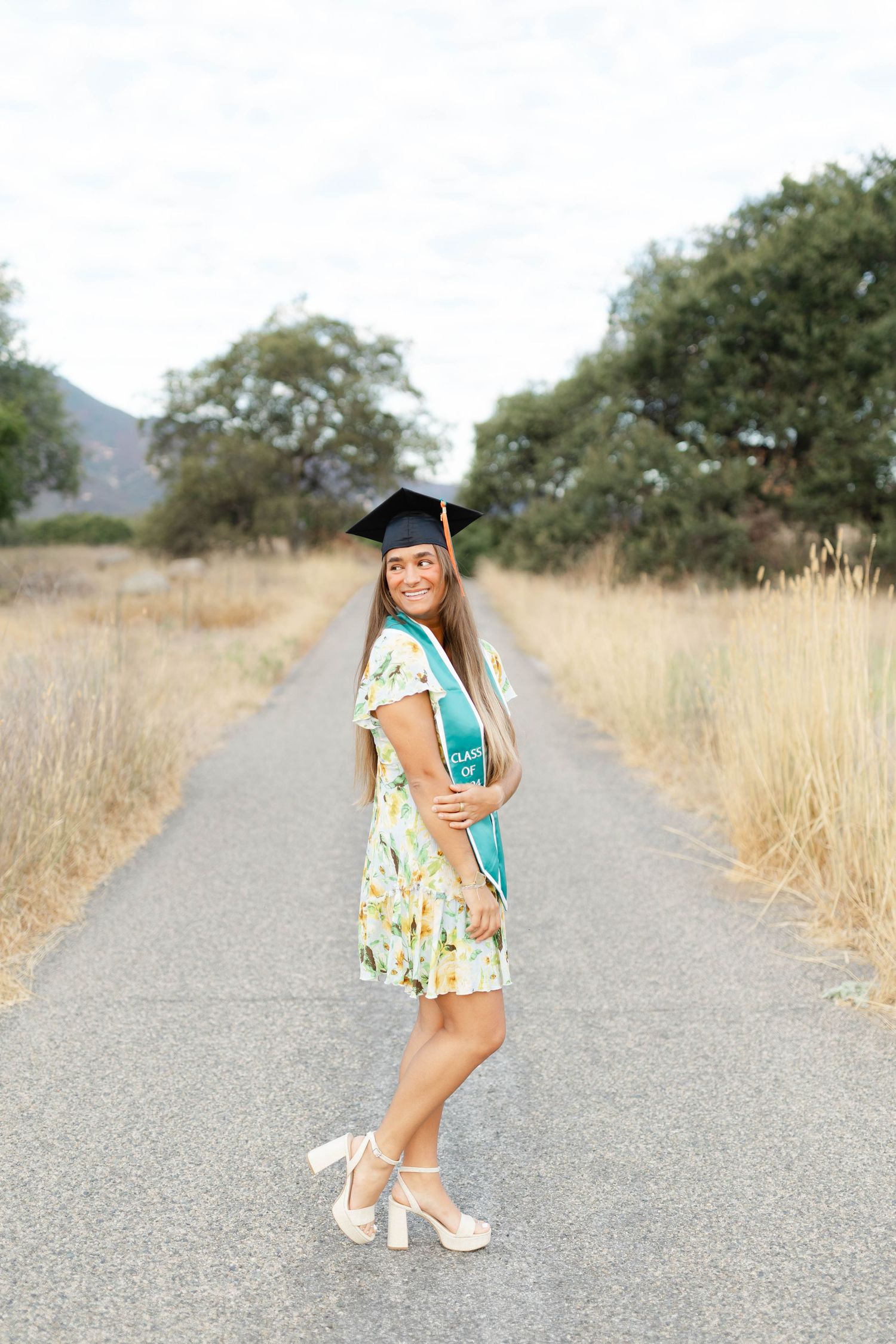 Graduate poses on a dirt path wearing a floral dress and graduation stole against a natural outdoor backdrop.