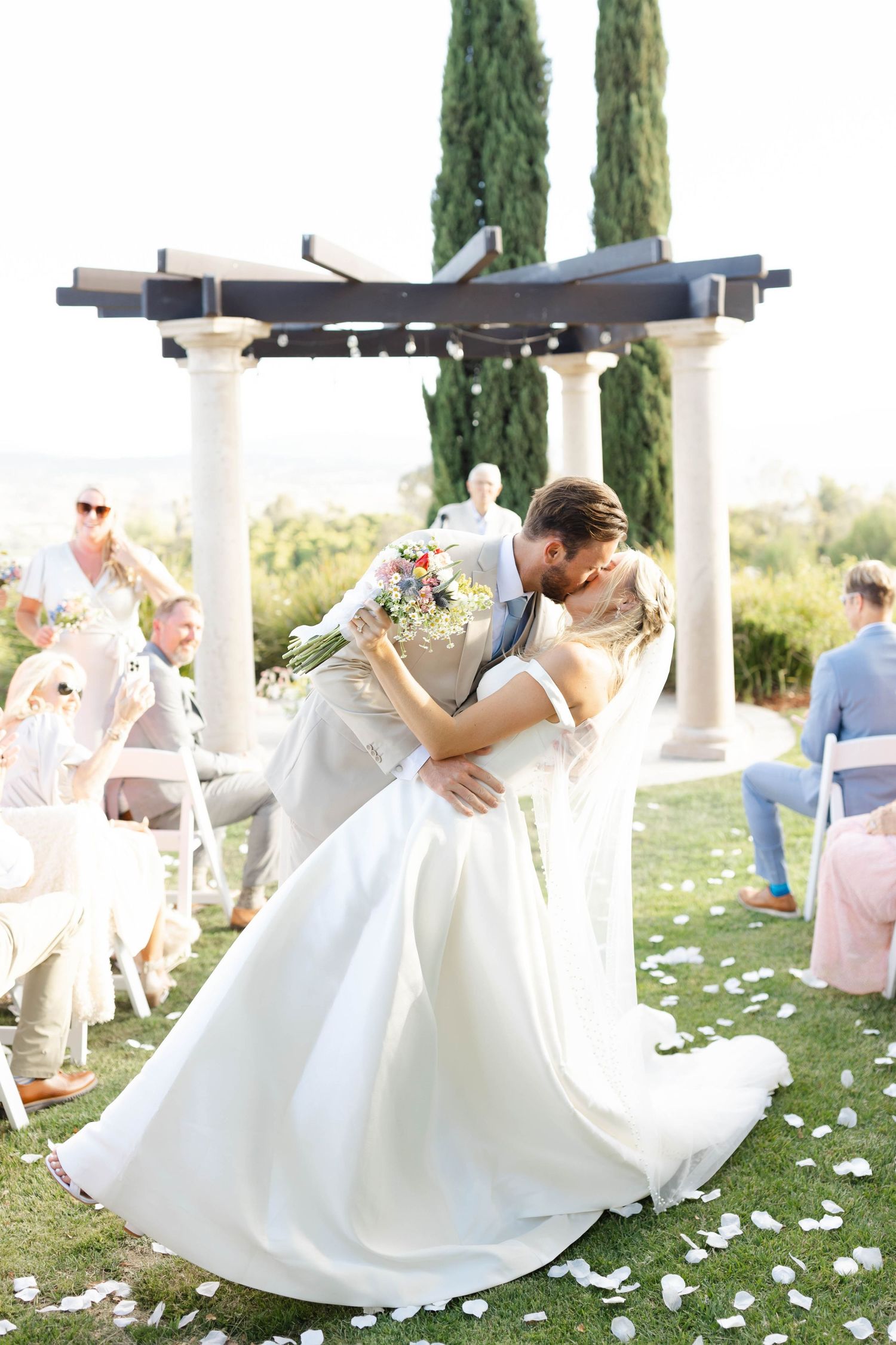 Dramatic dip kiss during wedding ceremony with guests and cypress trees in background.