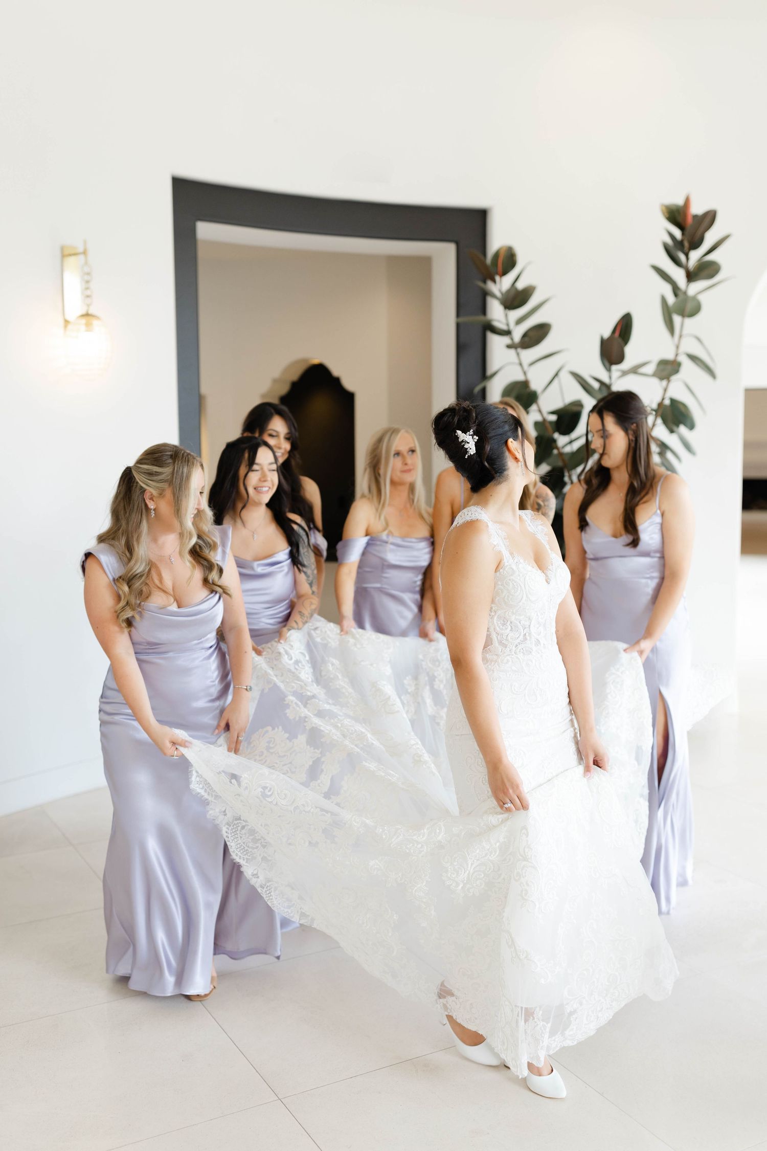 Bridesmaids in lavender dresses help adjust a flowing white wedding gown.