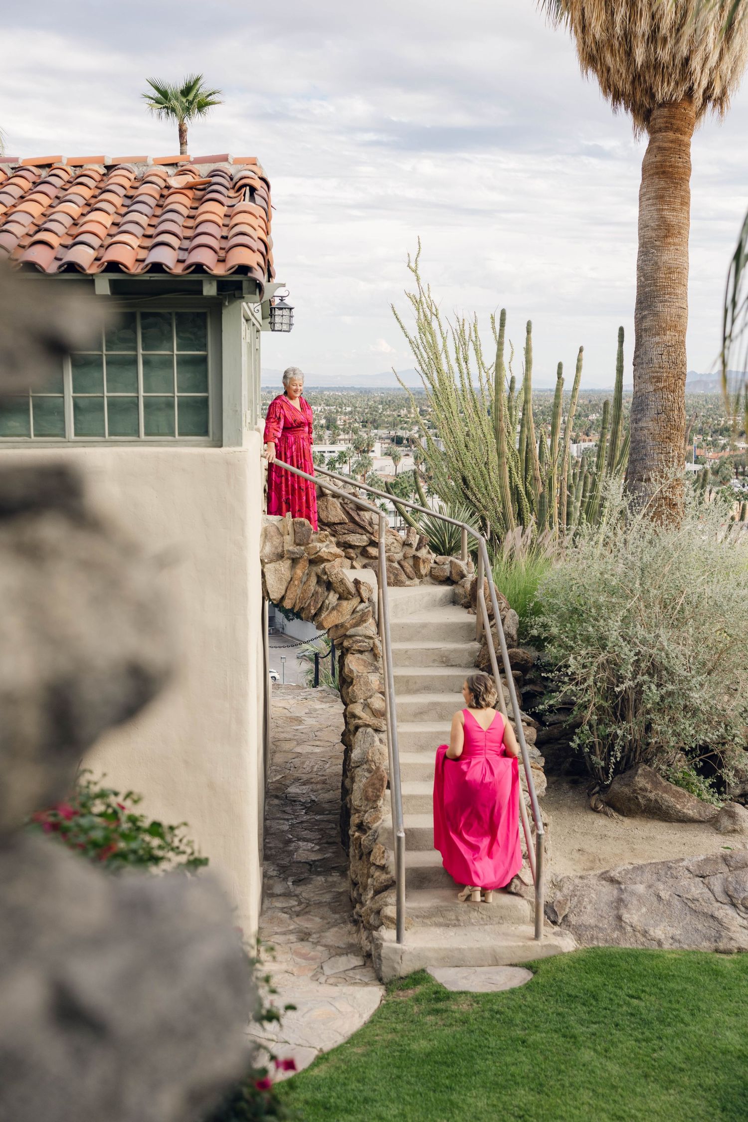 A red dress stands out against a desert landscape with palm trees and Spanish-style architecture in the background.