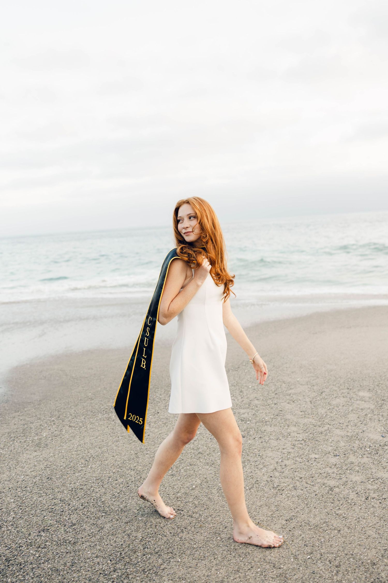 Young redhead woman in white dress holding graduation sash walks barefoot on beach with ocean backdrop.