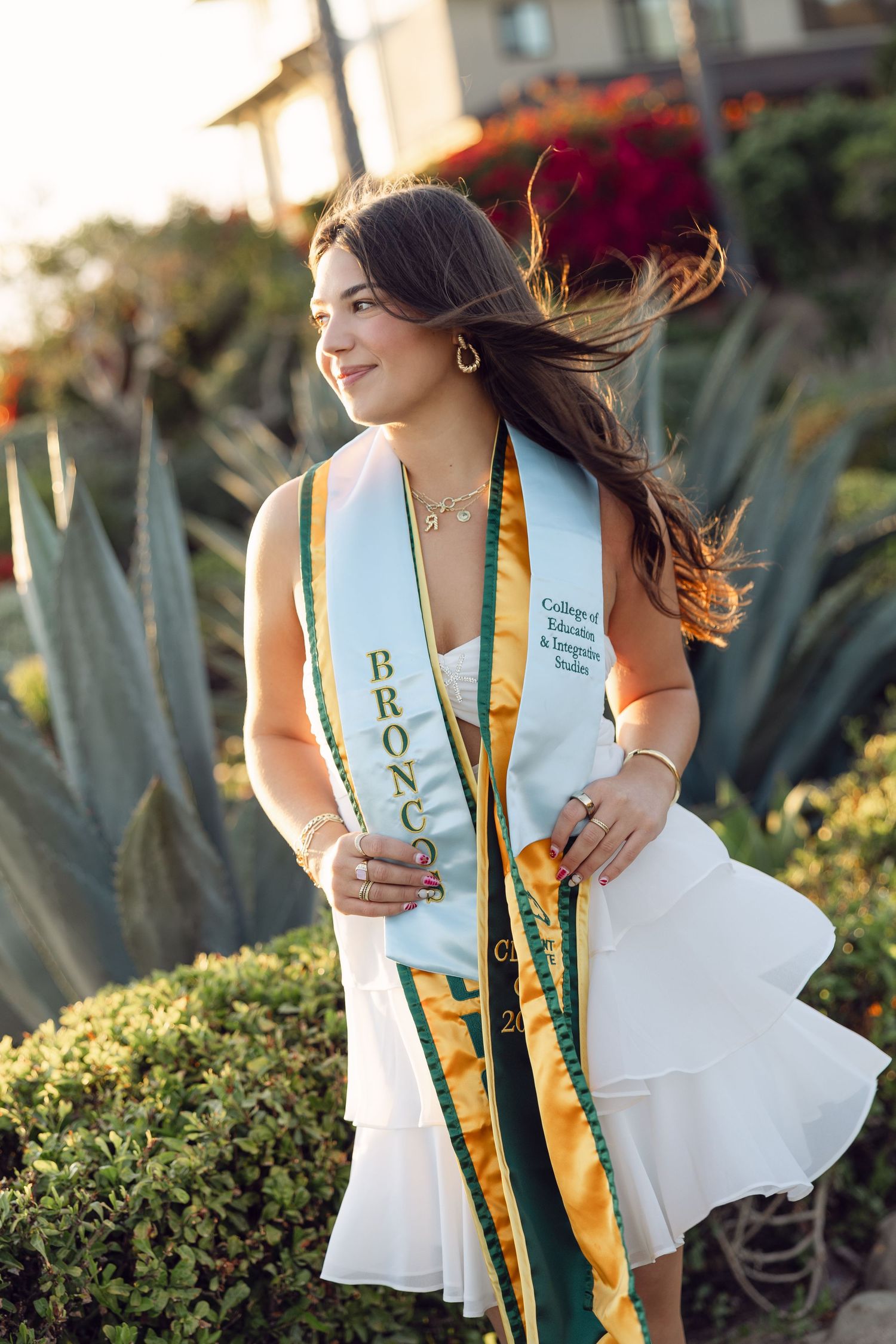 Young woman wearing green and gold Broncos graduation sash poses outdoors among agave plants at golden hour.