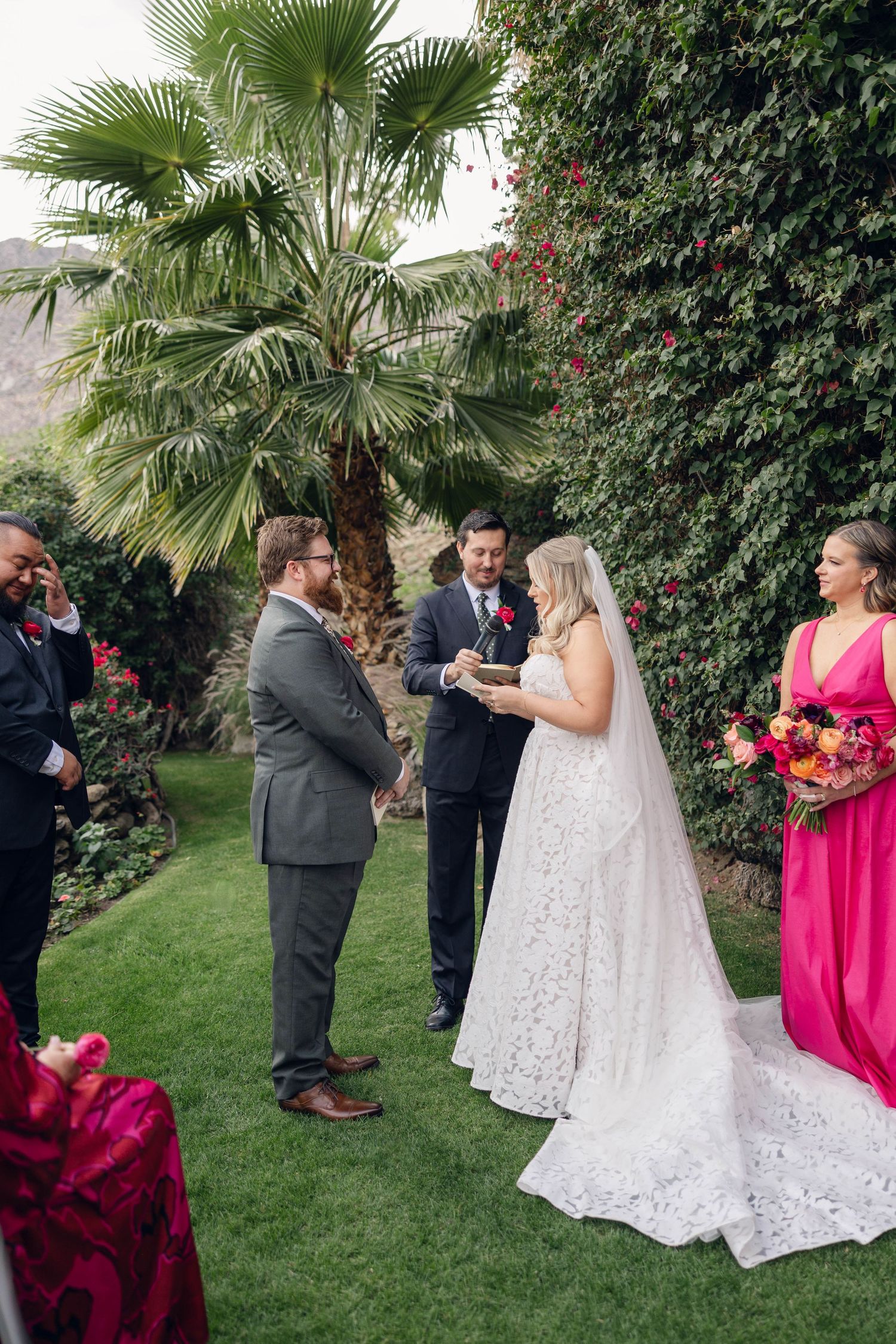 A romantic outdoor wedding ceremony moment with a bride in white lace and bridesmaids in bright pink dresses under palm trees.