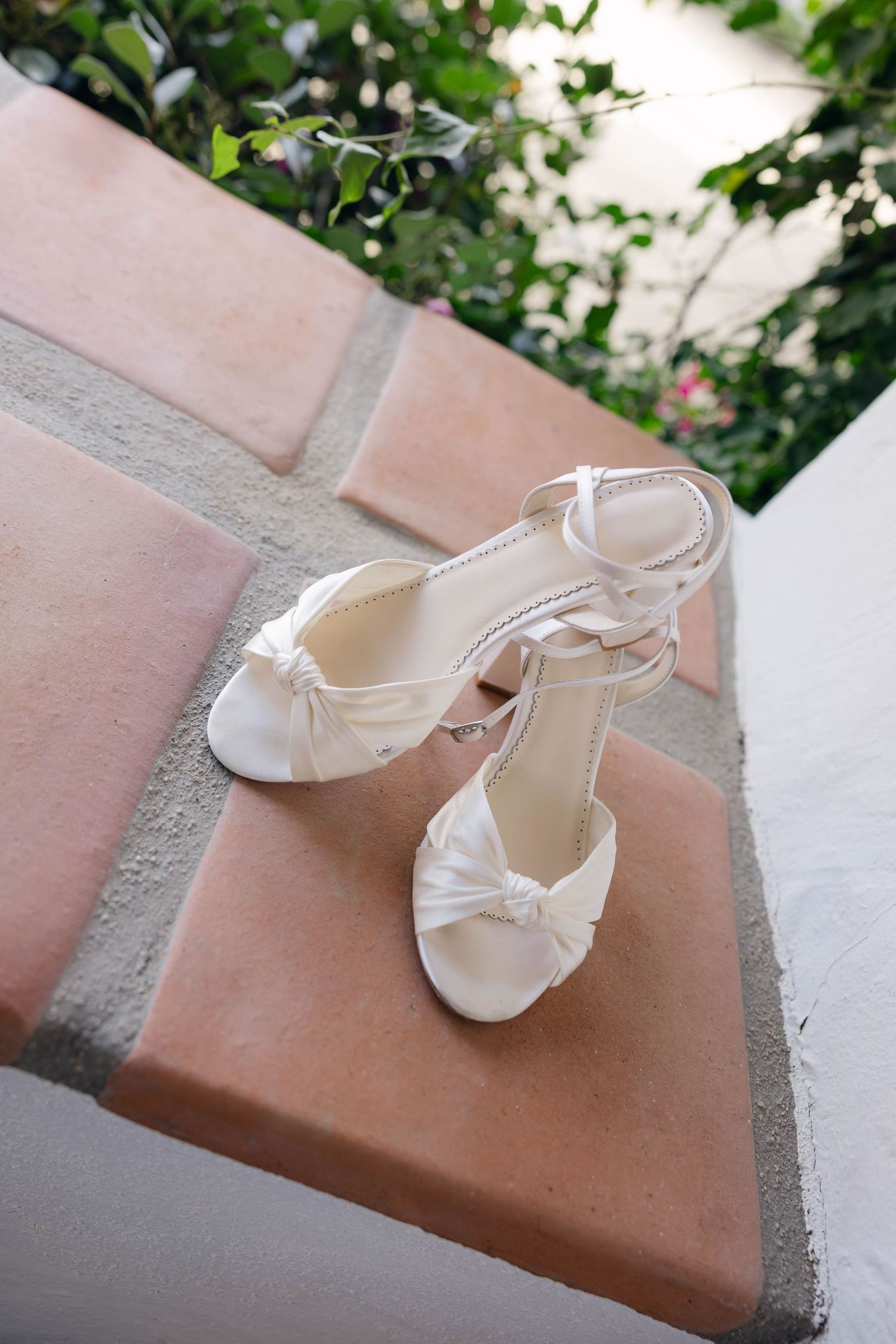 Elegant white satin bridal shoes with knotted details rest on a terracotta surface.