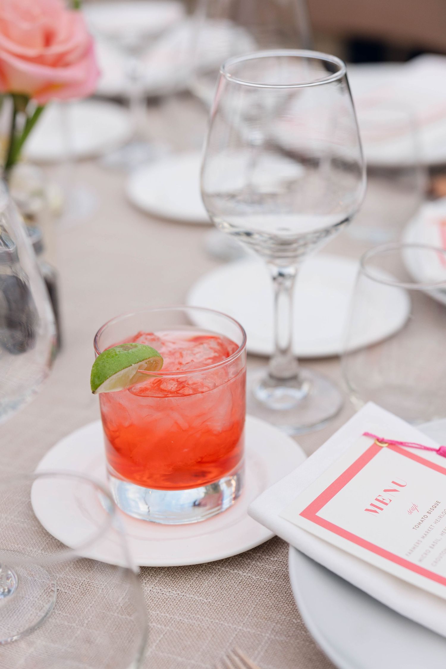 A red cocktail with lime garnish sits on a white plate next to an empty wine glass and menu card at an elegant table setting.