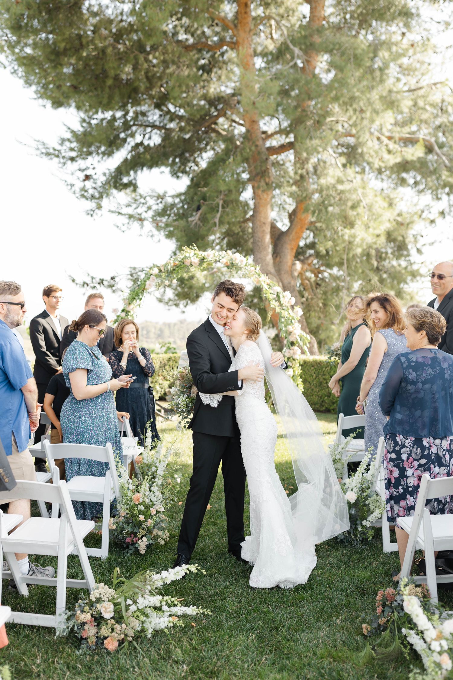 Guests mingle at an outdoor wedding ceremony under tall pine trees.