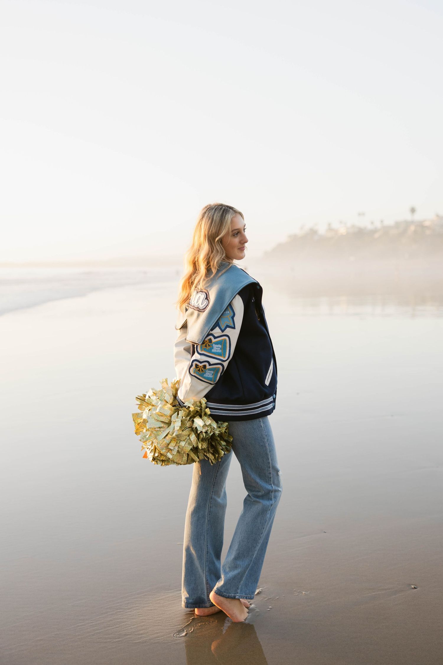 Person holding flowers walks along misty beach at sunset in casual denim outfit.