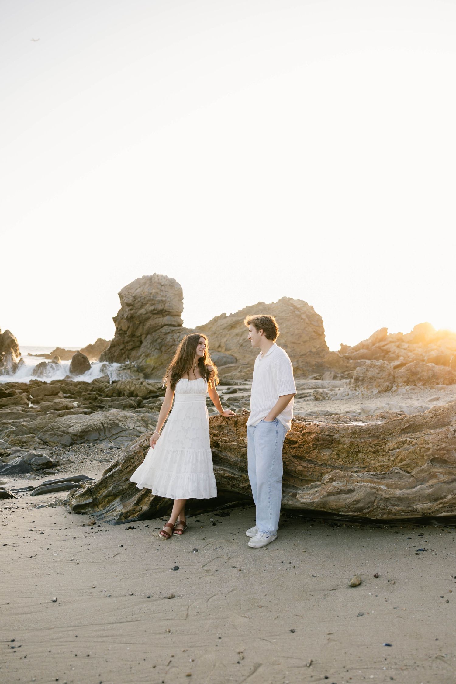 A couple in white outfits explores rocky beach formations at golden hour.
