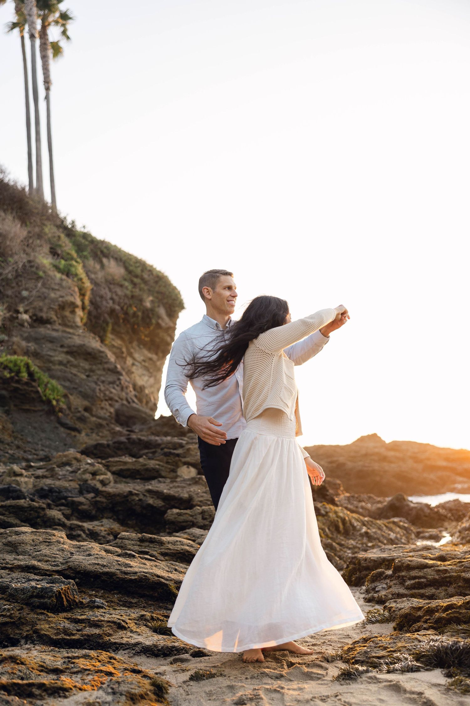 Couple dancing on rocky coastal cliffs at sunset with palm trees in background.