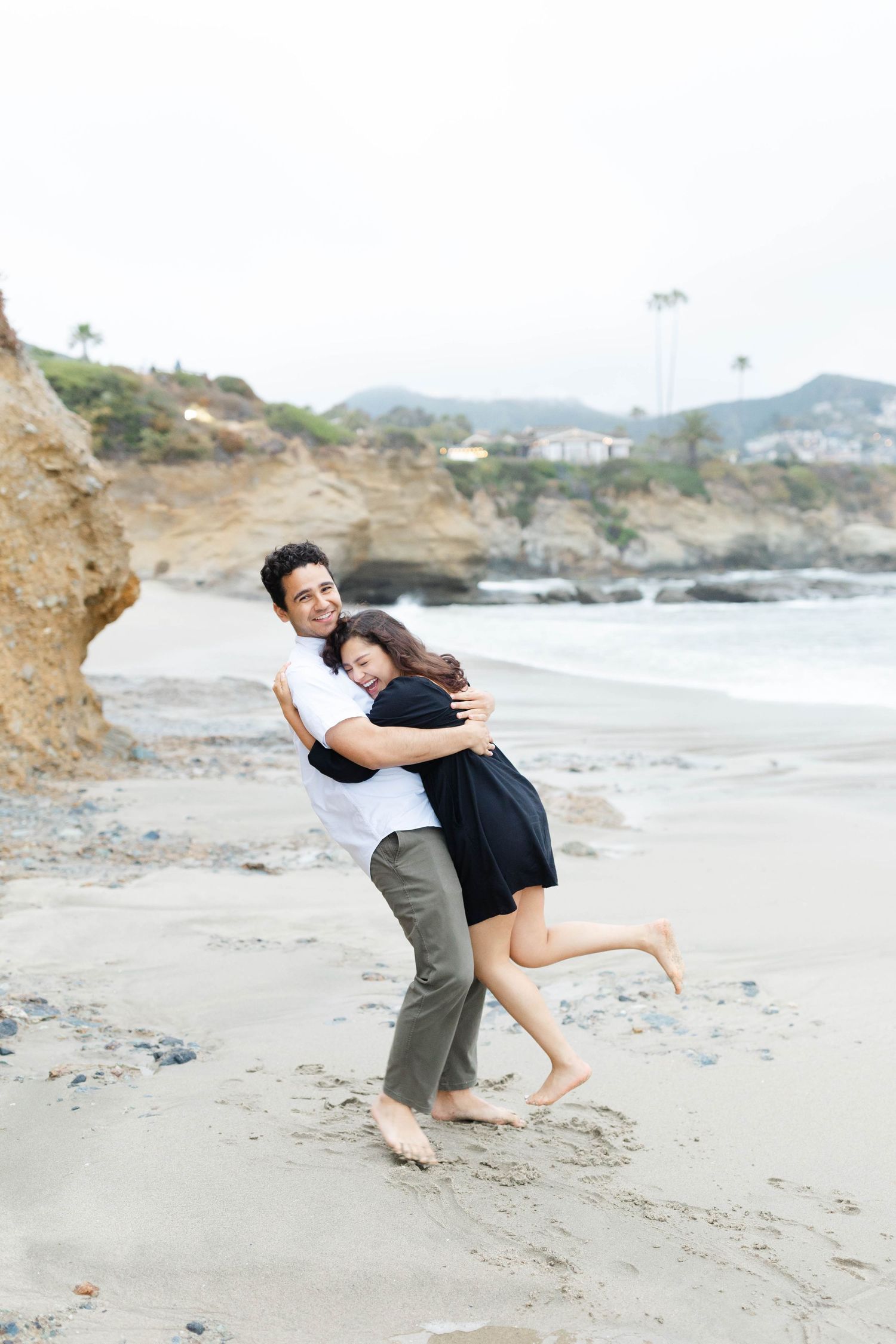 Romantic couple embracing on a scenic beach with coastal cliffs and waves in the background.