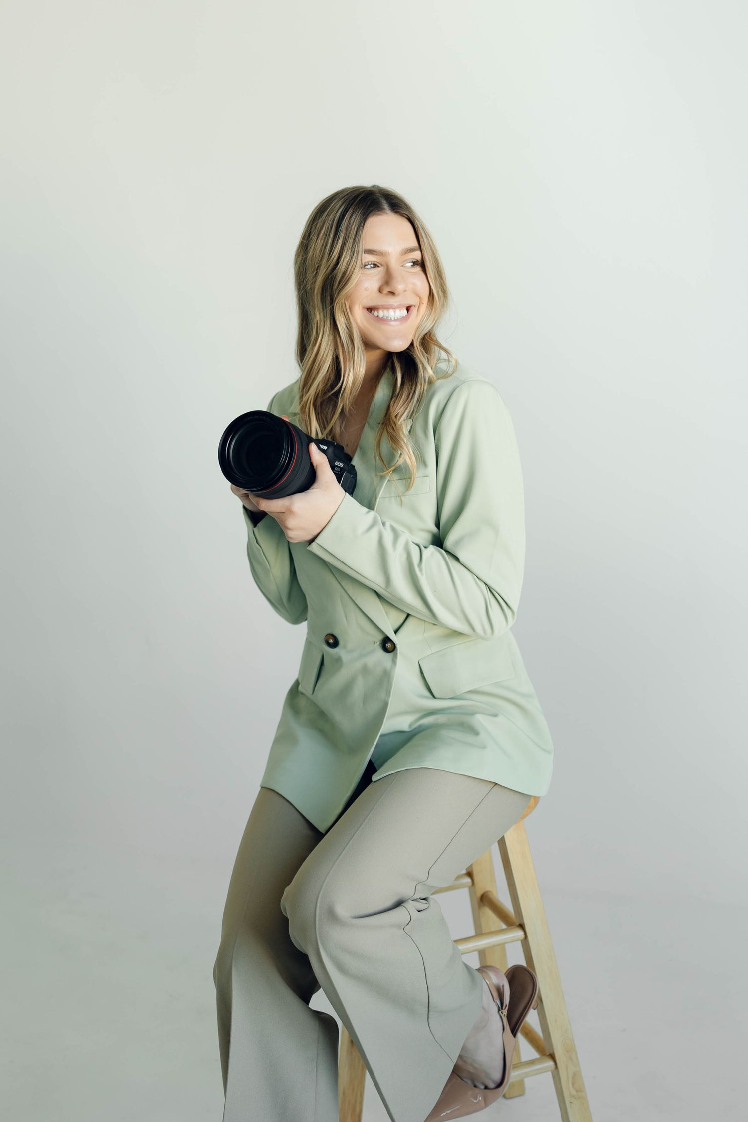 Professional model in mint green blazer poses playfully on wooden chair against white backdrop.