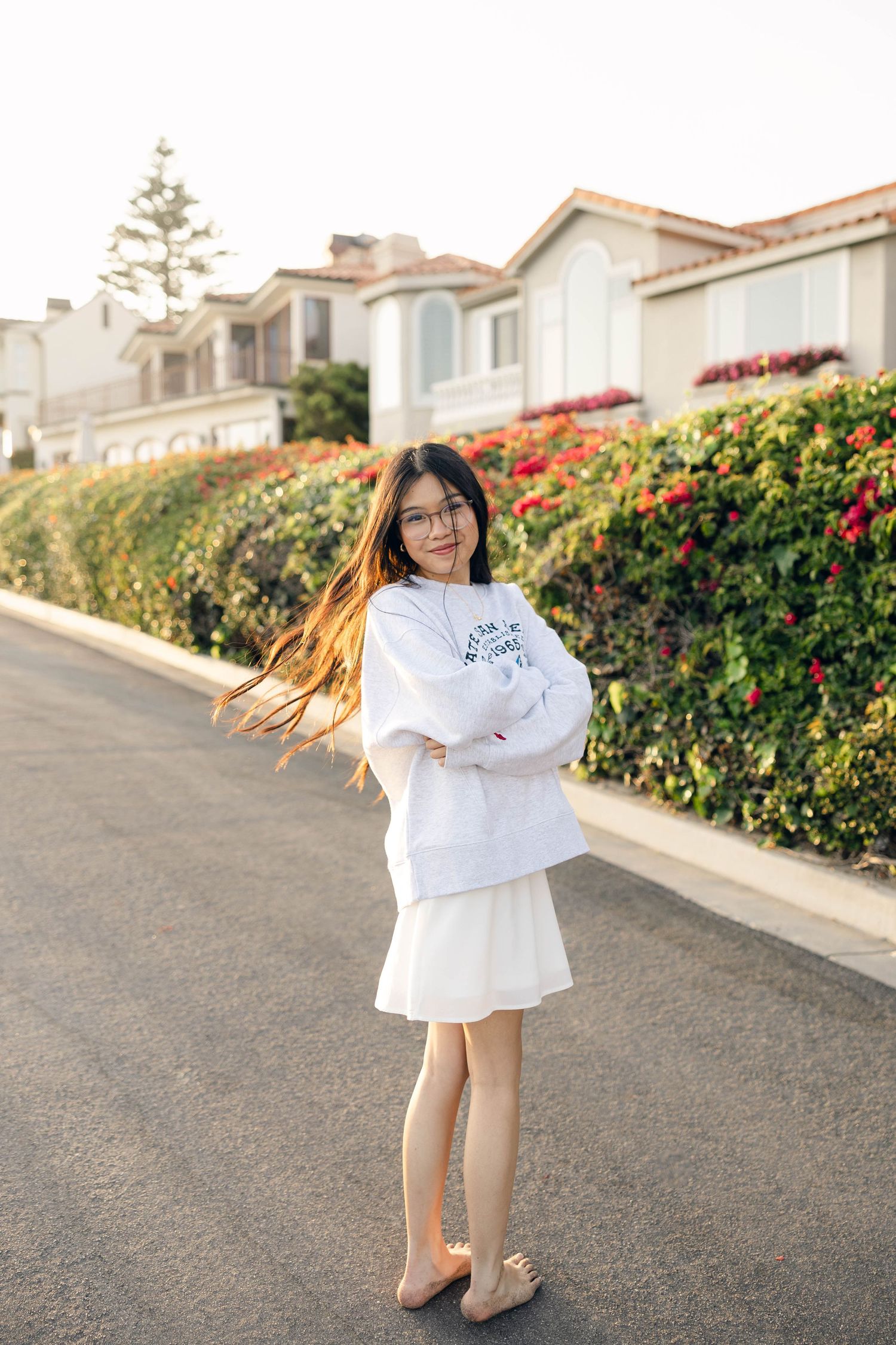 Person in white sweater twirls playfully on suburban street with flowering bushes.
