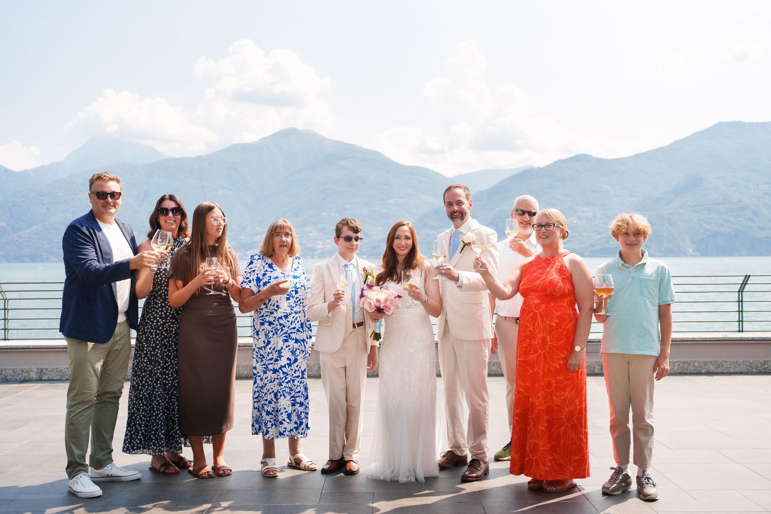 Wedding group photo with mountains and lake backdrop, guests toasting with champagne glasses outdoors.