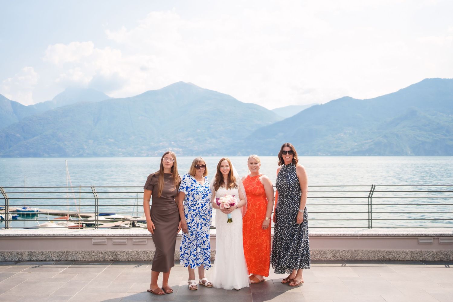 Five women posing together on a lakeside terrace with stunning mountain and water views in the background.