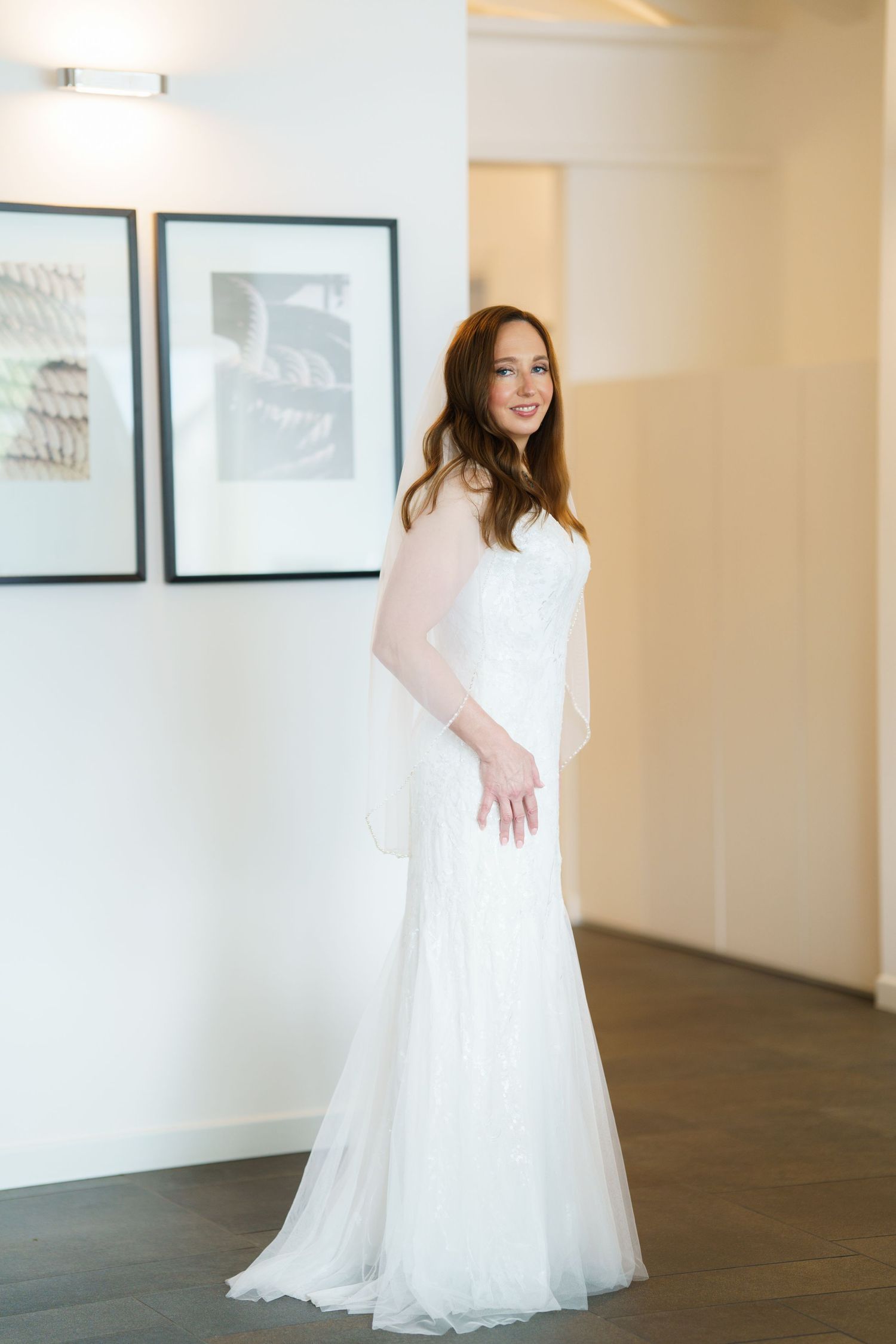 Smiling bride in elegant white tulle wedding gown posing in a bright room with framed artwork on the wall.