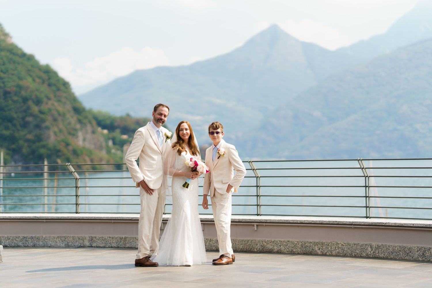 Bride in white gown with two guests in cream suits posing on terrace with mountain lake backdrop.