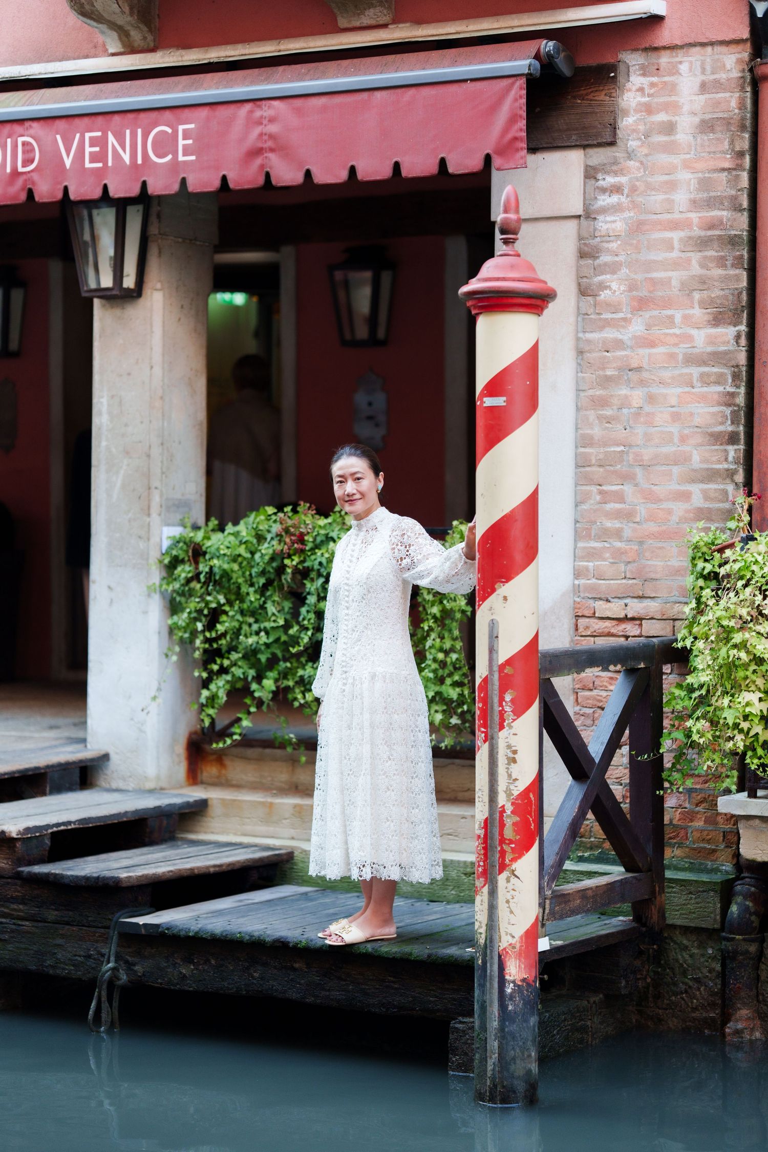 Woman in white lace dress posing by a striped mooring pole along a Venice canal.