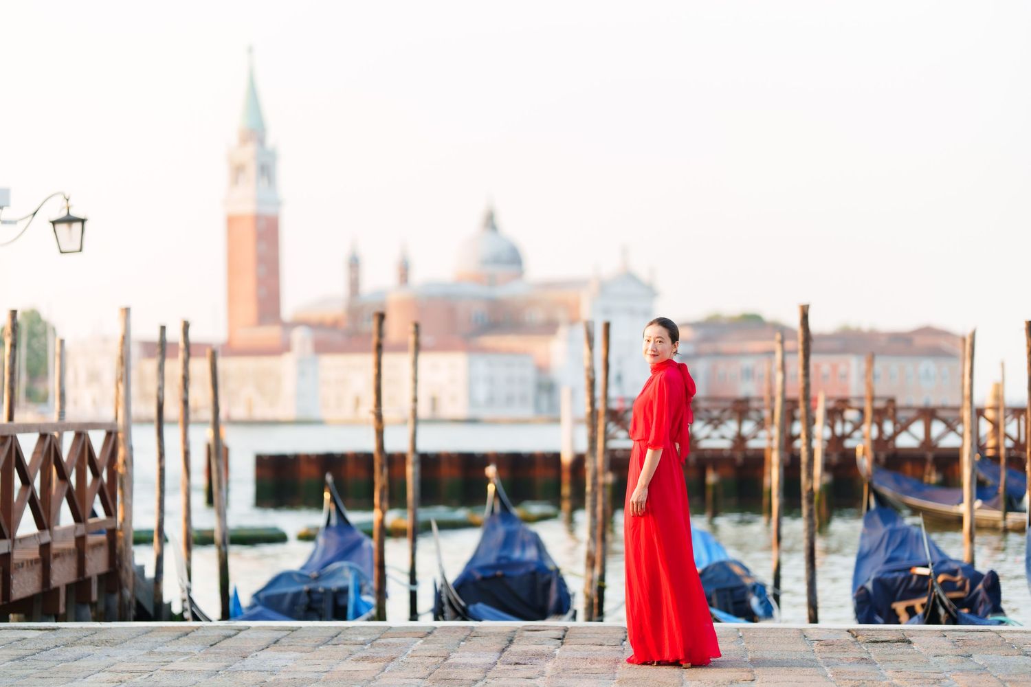 Woman in red dress standing by Venice canal with gondolas and San Giorgio Maggiore church in background.