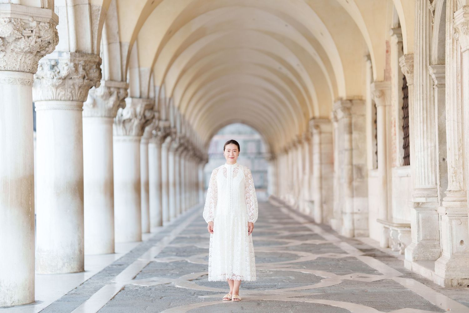 Woman in white dress walking through elegant Venetian arcade colonnade with arched ceilings in soft natural light.
