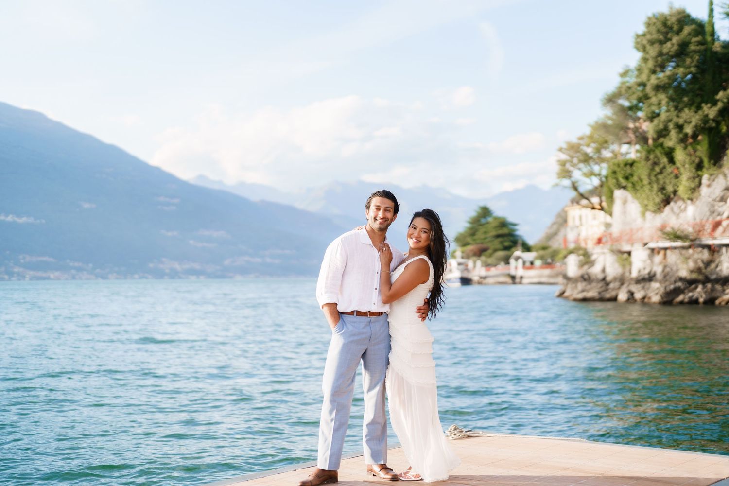 Couple posing on a lakeside dock with mountains and blue water in the background on a sunny day.