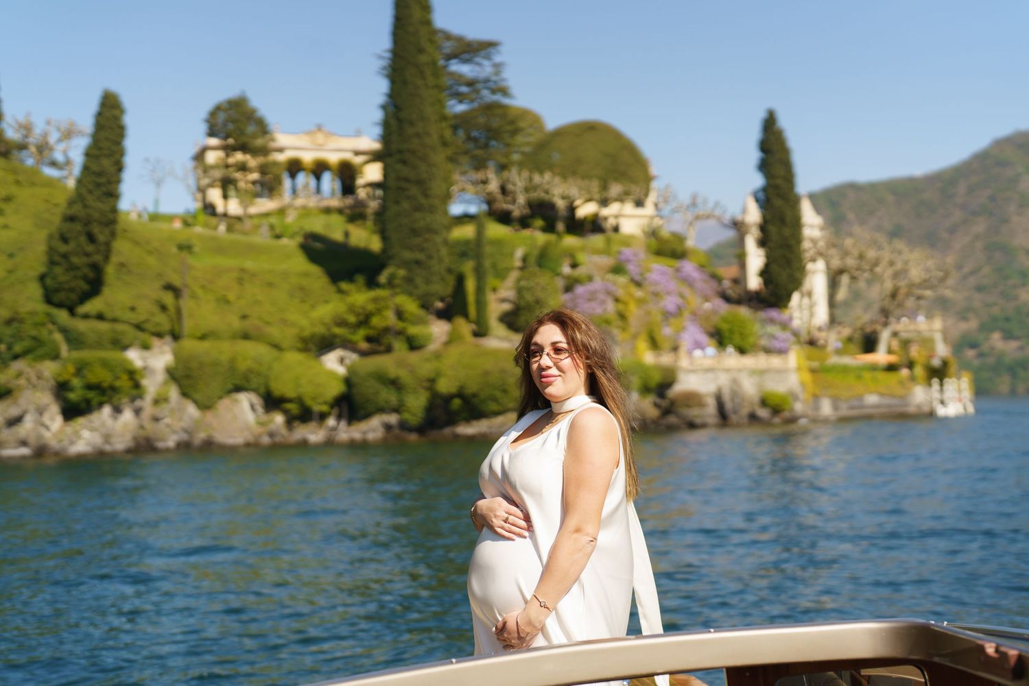 Pregnant woman in white dress poses on a boat with Villa del Balbianello and Lake Como in background.
