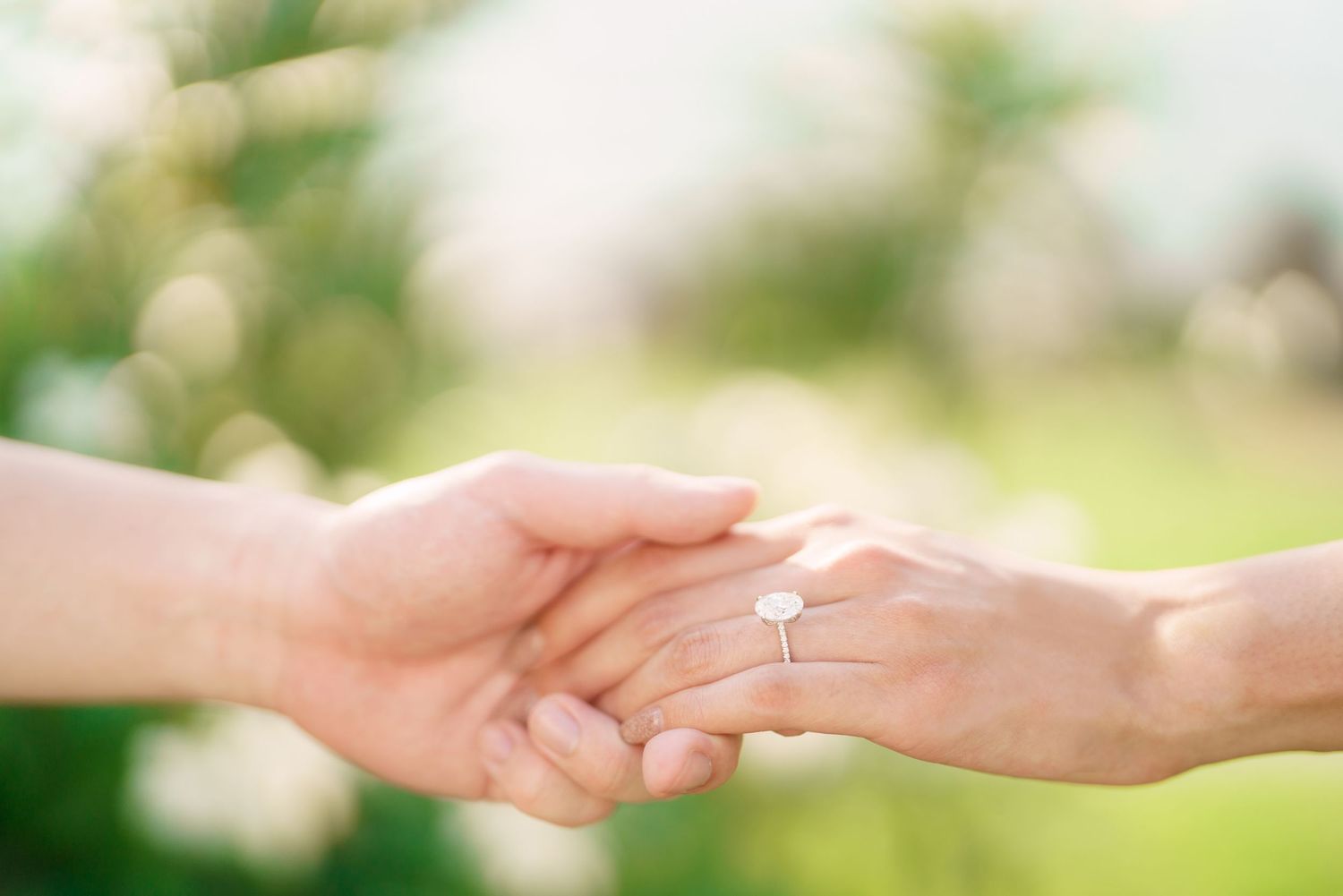 Two hands tenderly clasping together with an engagement ring sparkling in soft natural light against a blurred green background.