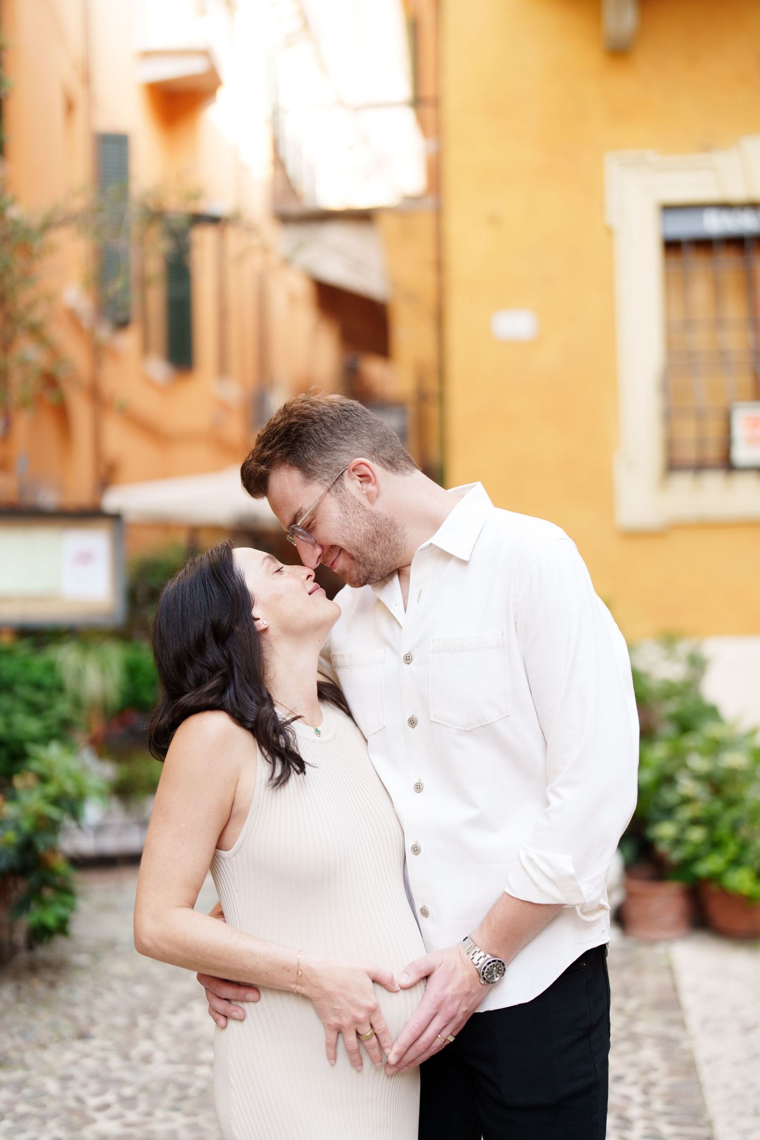 A romantic couple shares an intimate kiss in front of a warm yellow stucco wall on a cobblestone street.