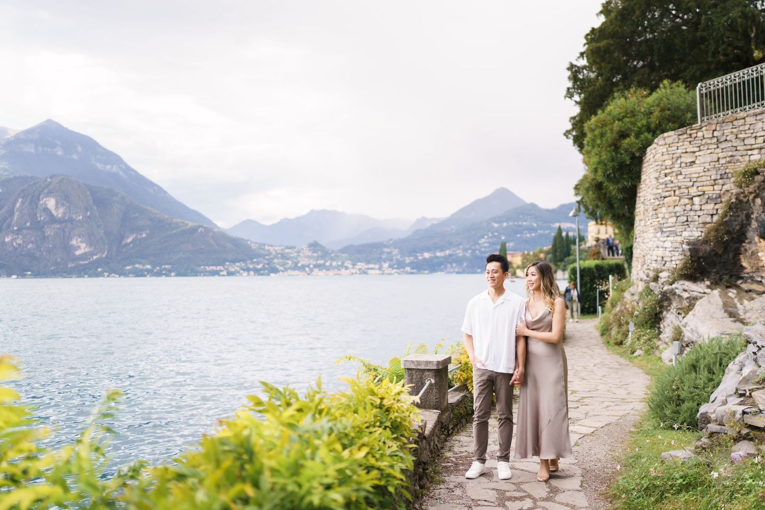 Couple walking along scenic lakeside path with mountain views in Lake Como, Italy.